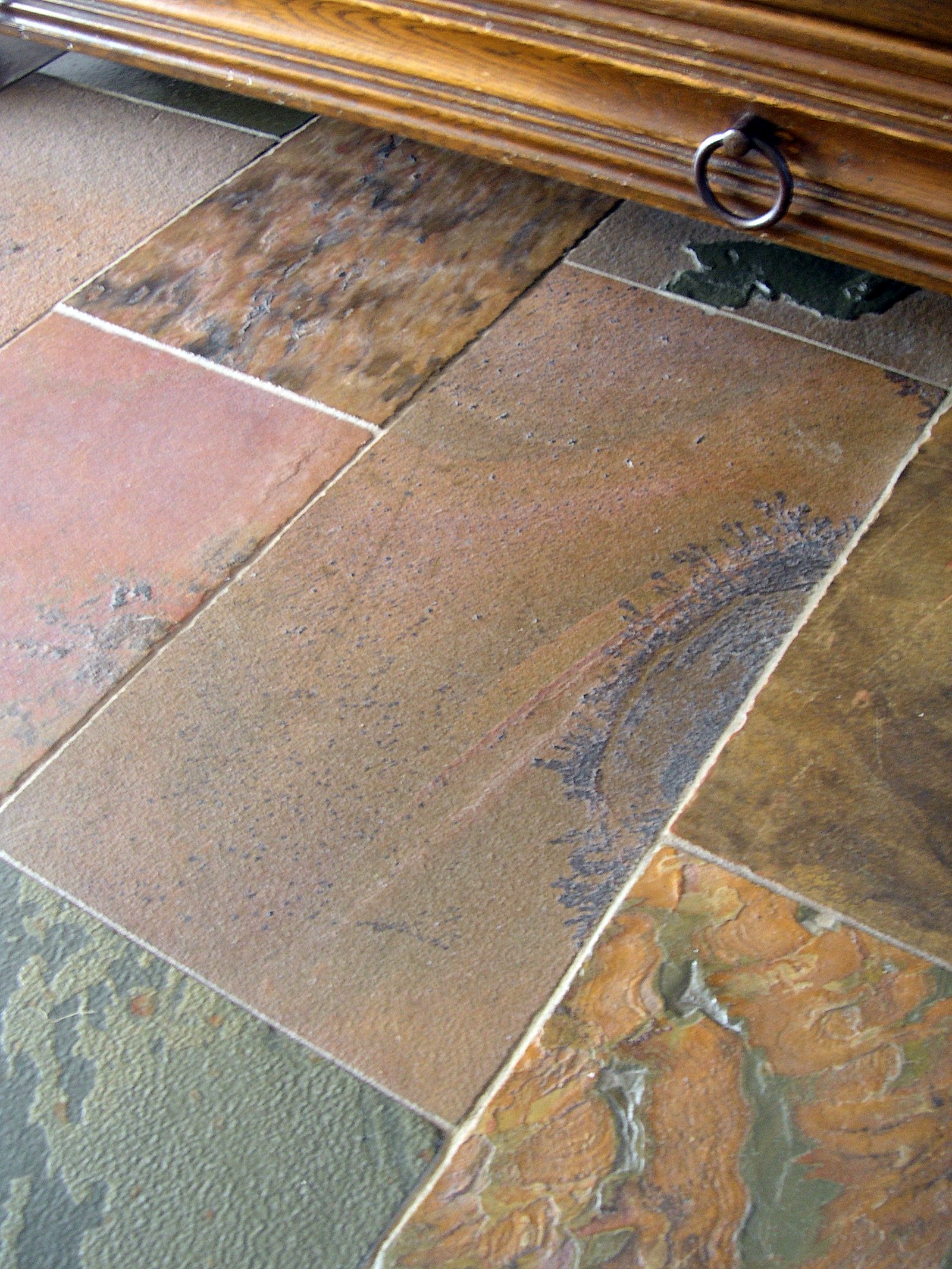 Fragment podłogi wyłożonej płytami z łupka afrykańskiego o zróżnicowanej kolorystyce i fakturze, widoczny fragment drewnianej komody z metalowym uchwytem.