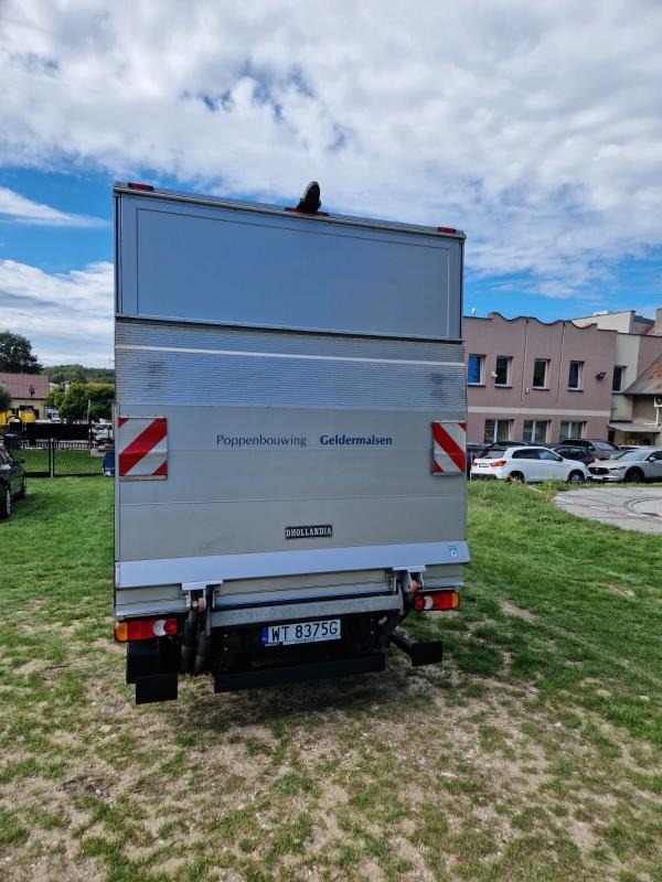 Tył srebrnego busa dostawczego z napisem 'Poppenbouwing Geldermalsen' na tle zieleni i budynków, widoczna tablica rejestracyjna WT 8375G.