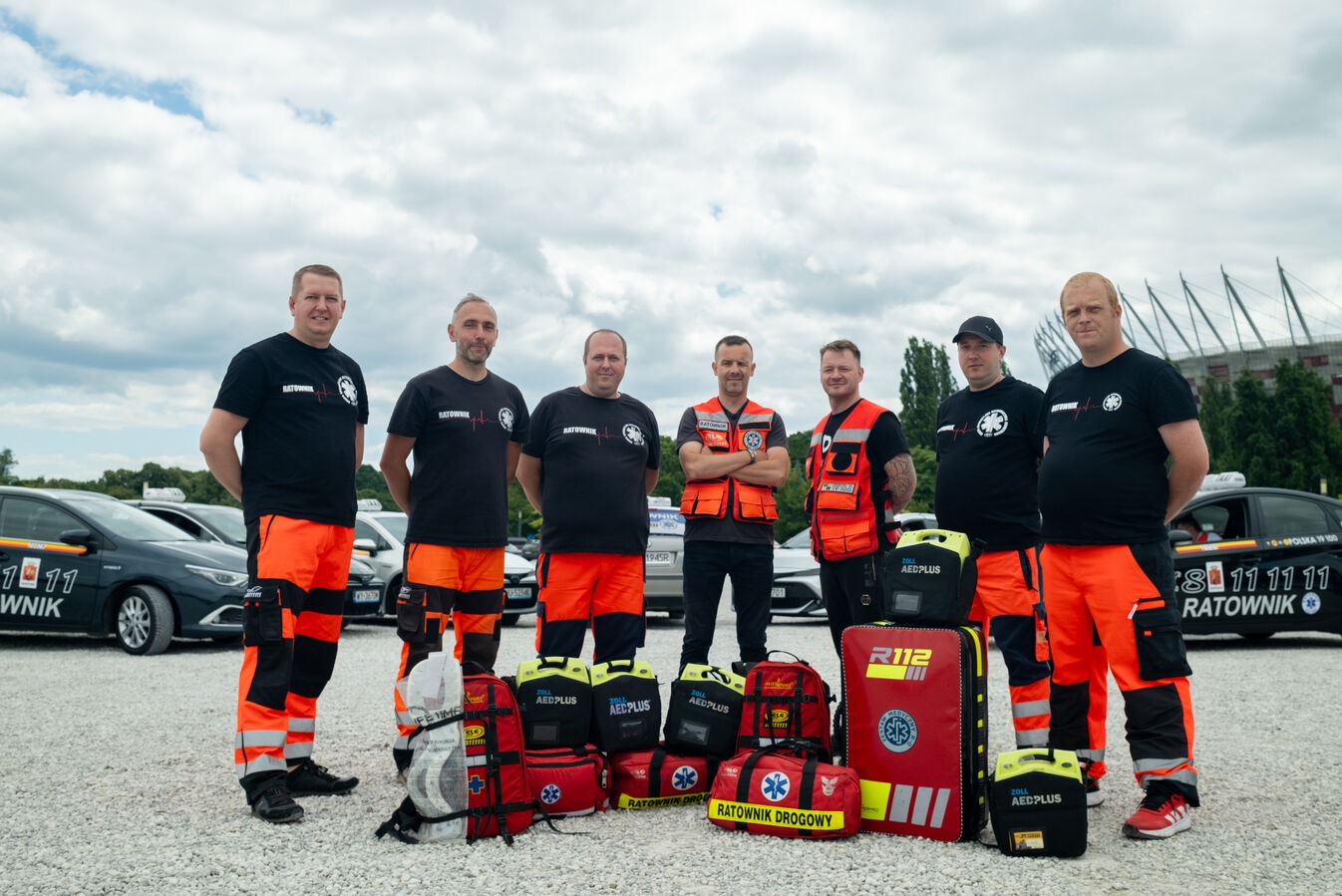 Grupa ratowników drogowych w czarno-pomarańczowych uniformach, stojąca przed samochodami z napisem 'Ratownik' i torbami medycznymi, na tle Stadionu Narodowego.