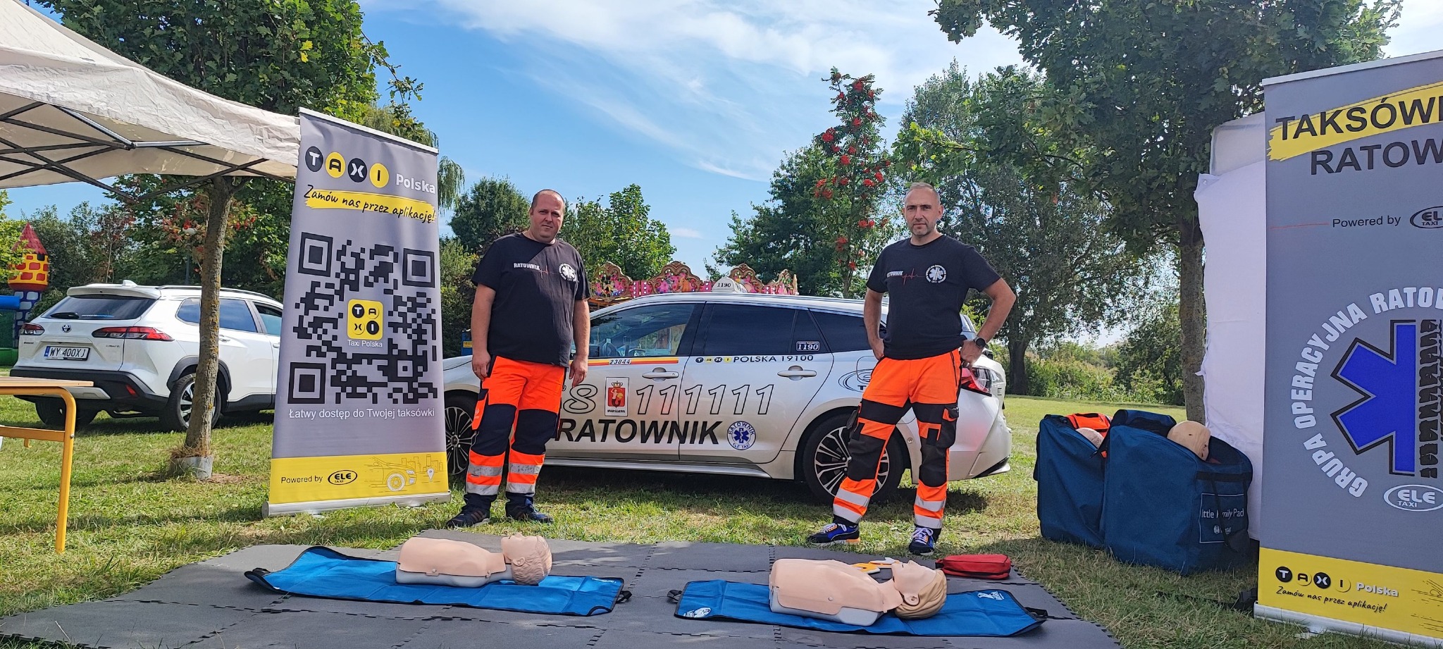 Dwóch ratowników medycznych w pomarańczowych kombinezonach prezentuje manekiny do ćwiczeń resuscytacji na trawie, w tle samochód Taxi Polska z numerem alarmowym i banery reklamowe.