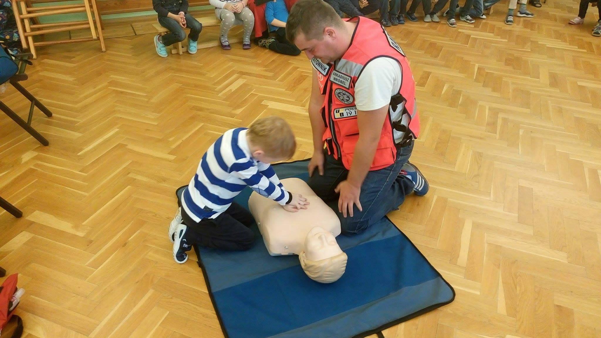 Instruktor w pomarańczowej kamizelce ratowniczej uczy małego chłopca uciskania klatki piersiowej na fantomie na tle parkietu i grupy osób.
