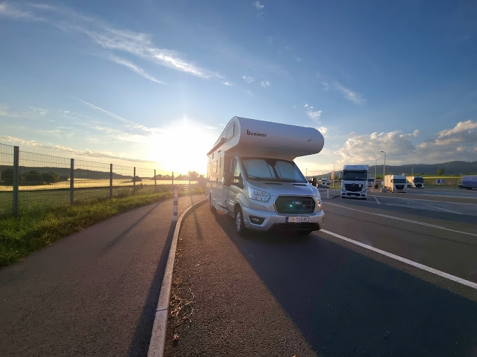 Srebrny kamper Benimar zaparkowany na parkingu przy drodze, z widokiem na zachodzące słońce i inne pojazdy w tle.