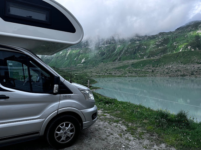 Srebrny kamper Ford Transit 160 zaparkowany nad brzegiem jeziora z widokiem na góry spowite mgłą.