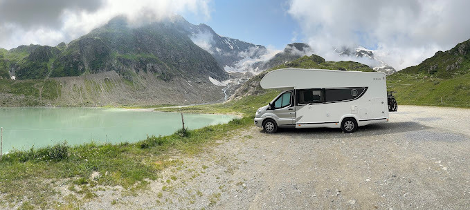 Kempingowy samochód na tle górskiego jeziora o turkusowej wodzie i ośnieżonych szczytów.