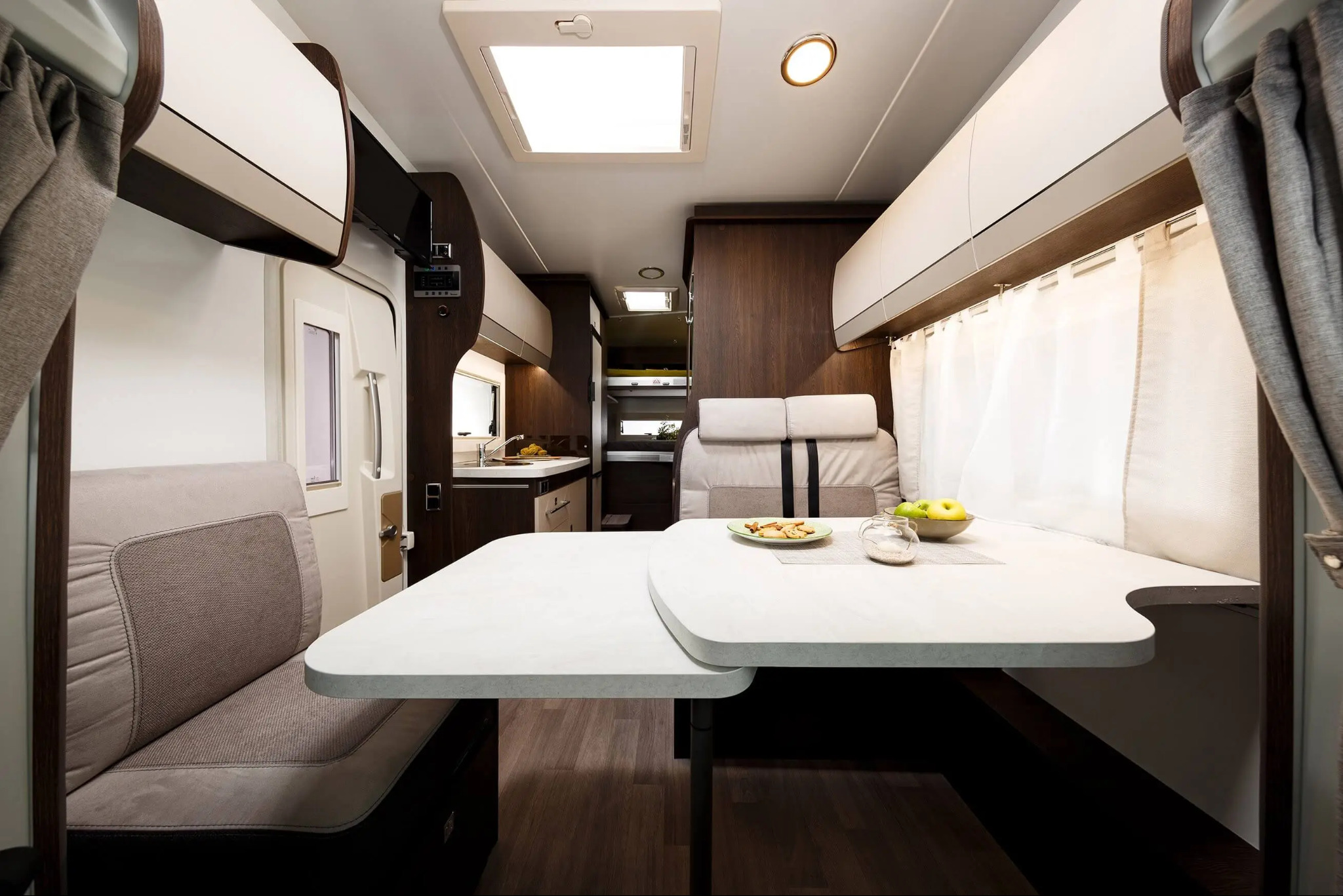 Wnętrze kampera z rozkładanym stolikiem, kanapą i aneksem kuchennym; na stole owoce i ciastka.