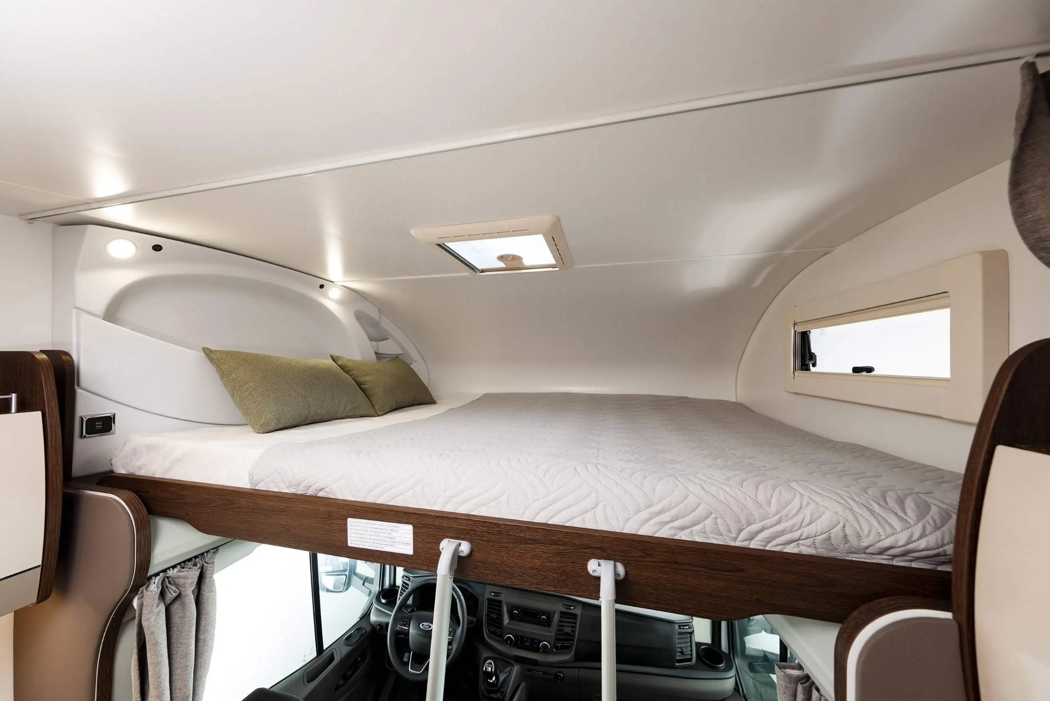 Wnętrze kampera z łóżkiem nad kabiną kierowcy we Wrocławiu, widoczne zielone poduszki i sterownica pojazdu.
