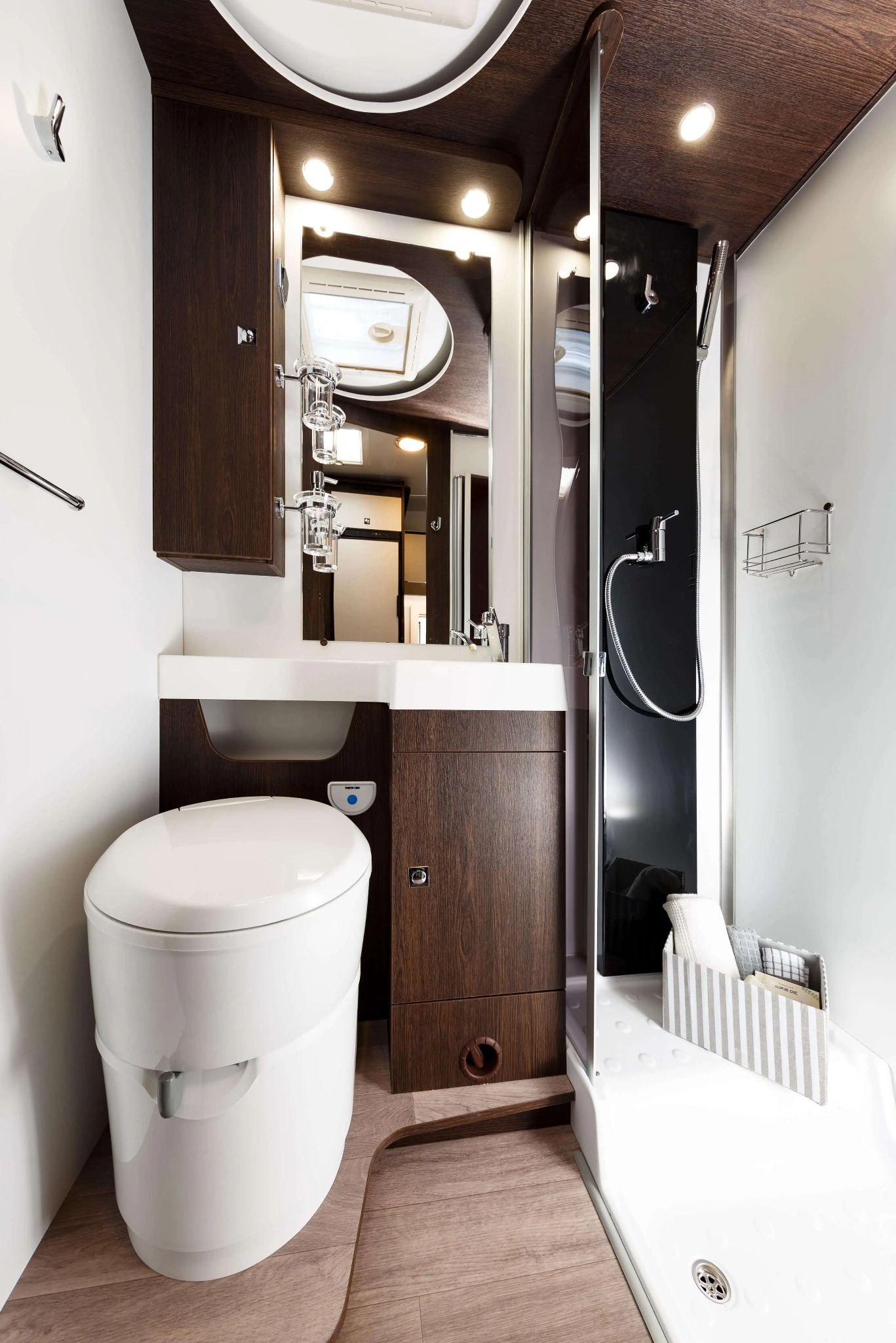 Kompaktowa łazienka w kamperze z toaletą, umywalką, szafkami z ciemnego drewna i kabiną prysznicową z czarnym panelem.