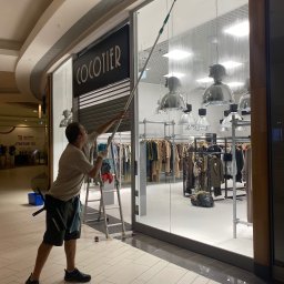 As-po - Mężczyzna myje wysoką witrynę sklepu odzieżowego 'COCOTIER' w galerii handlowej, używając długiego kija z gumową ściągaczką, drabina obok.