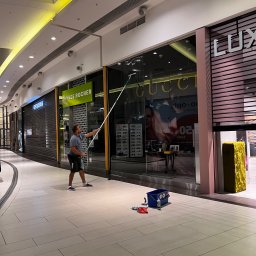 As-po - Pracownik w szarej koszulce czyści wysokie okno wystawowe sklepu Gucci w galerii handlowej za pomocą długiego mopa, obok stoi niebieskie wiadro z akcesoriami.