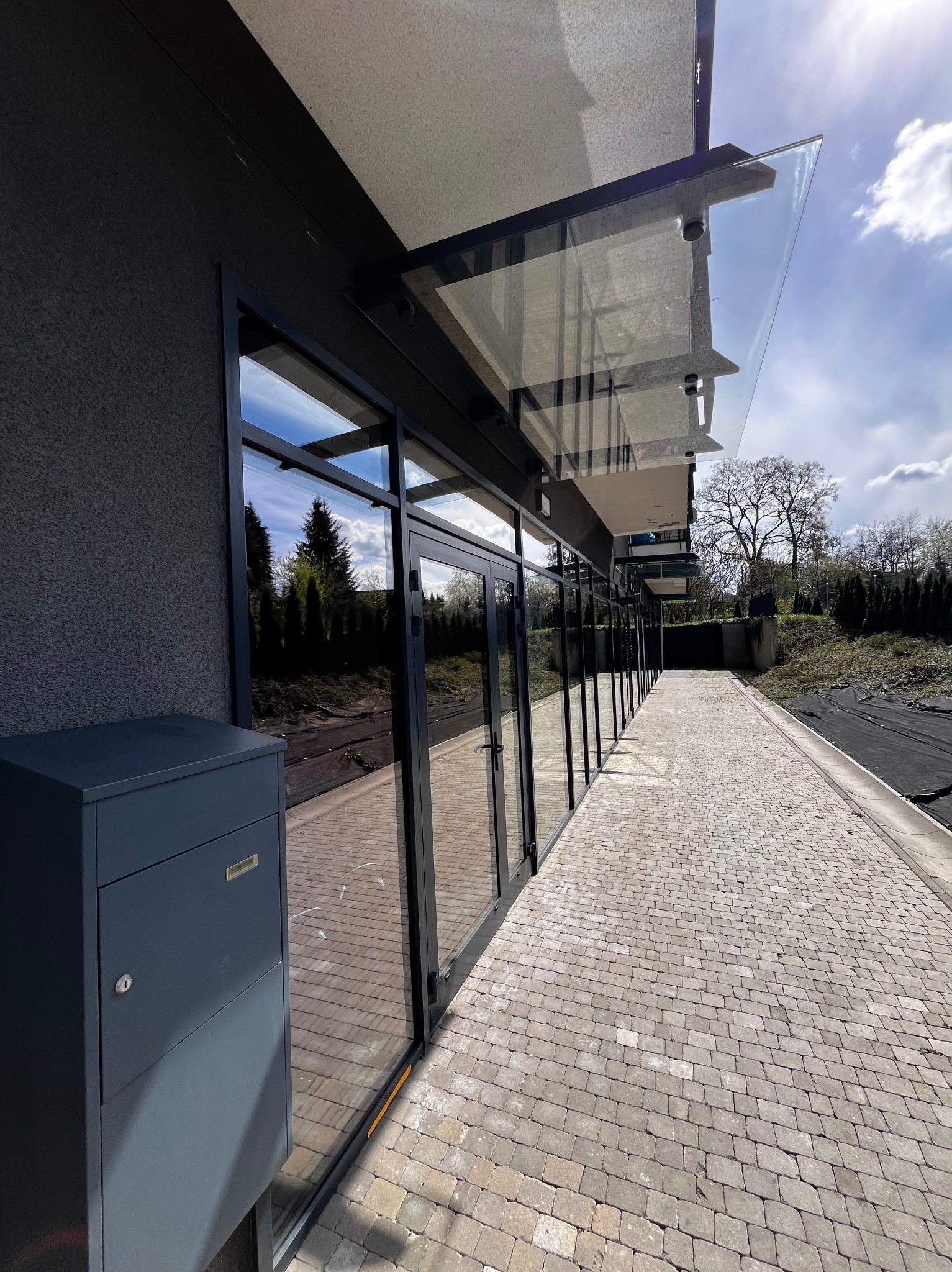 Szereg czarnych okien biurowych z szklanymi daszkami nad wejściami, odbijających niebo i drzewa, obok brukowany chodnik i szara skrzynka pocztowa.