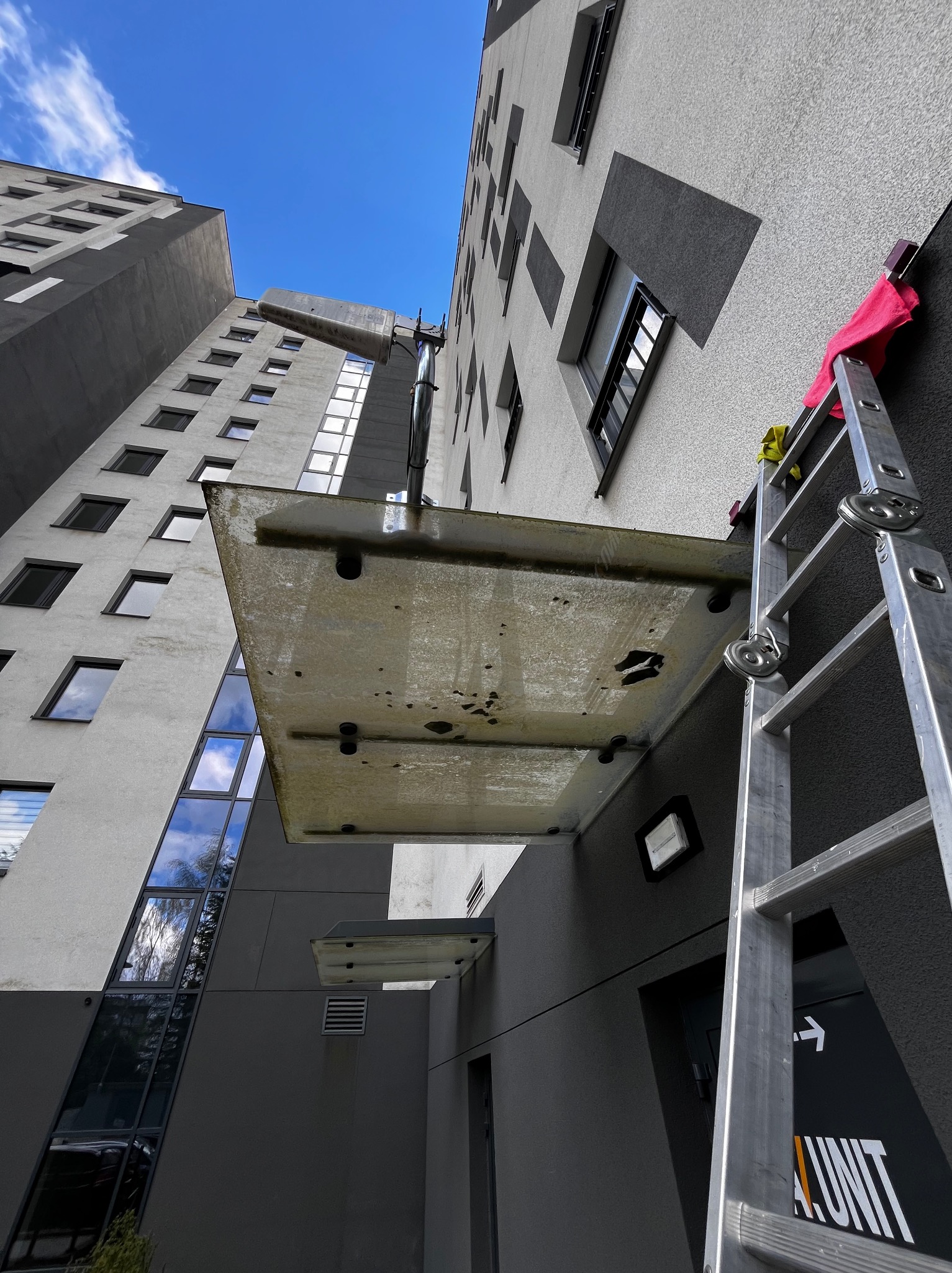 Wysoki budynek z widoczną elewacją i oknami, drabina oparta o ścianę z czerwoną szmatką, element konstrukcyjny nad wejściem do budynku.