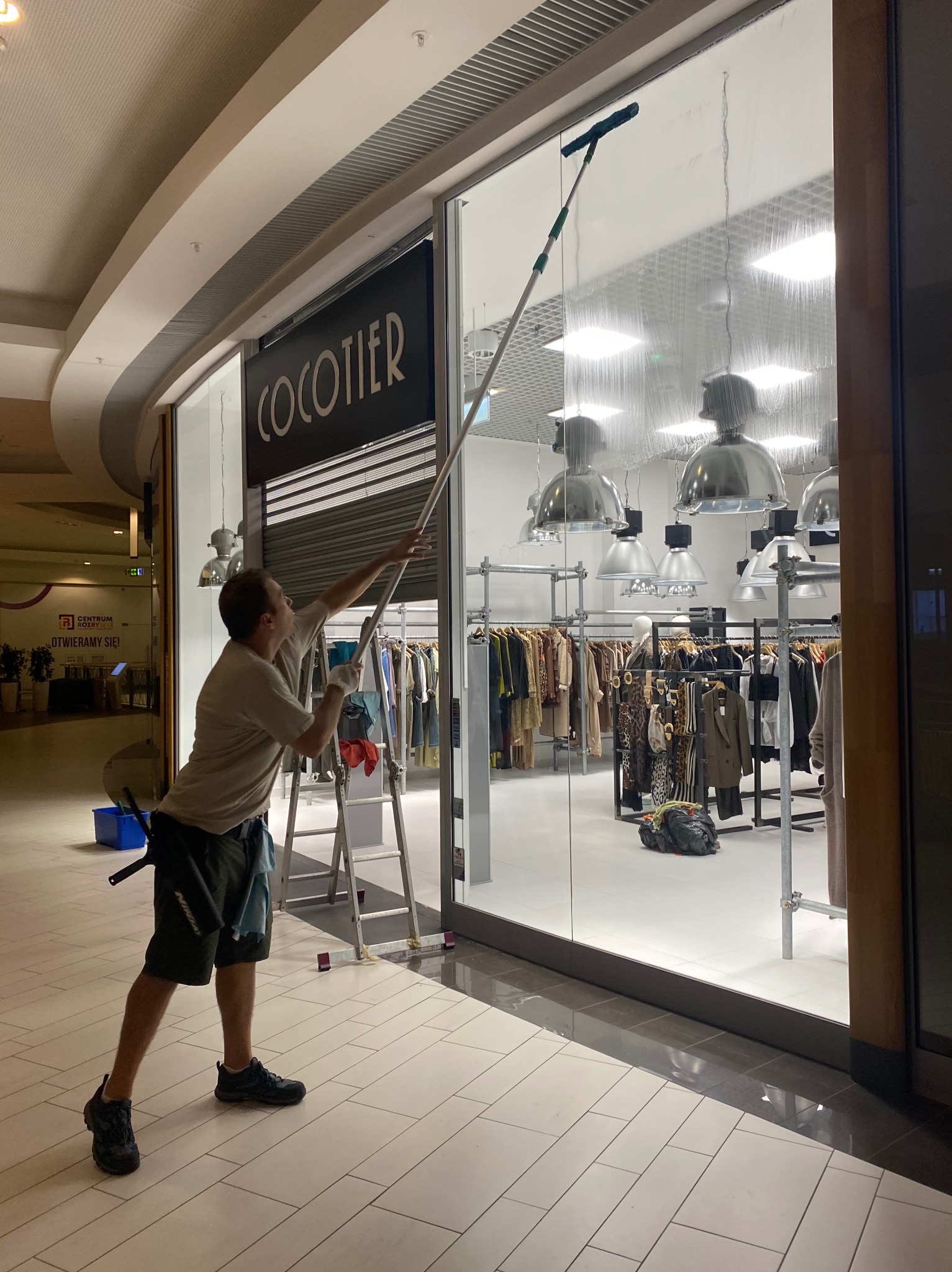 Mężczyzna myje wysoką witrynę sklepu odzieżowego 'COCOTIER' w galerii handlowej, używając długiego kija z gumową ściągaczką, drabina obok.
