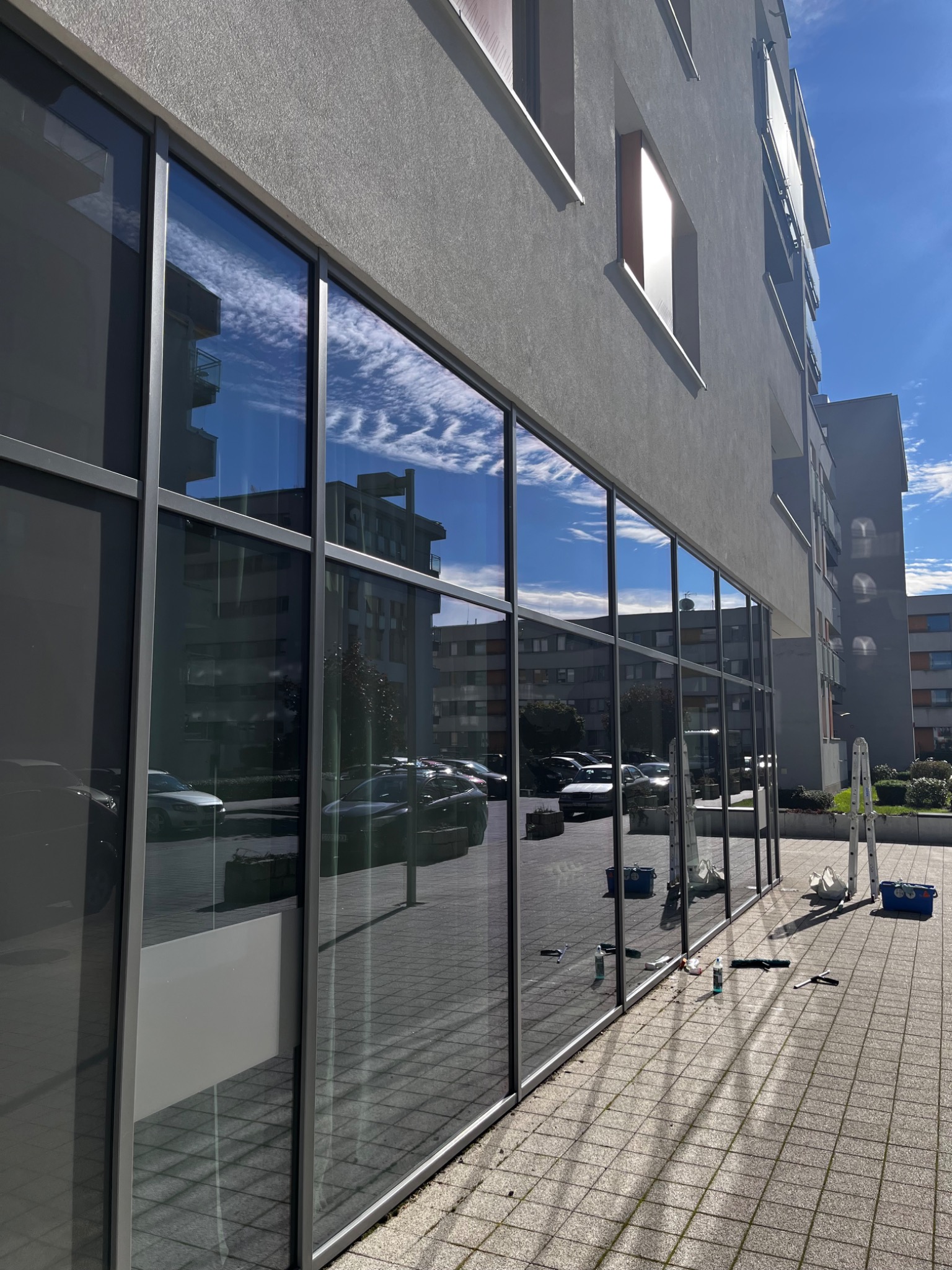 Duże okna biurowe z aluminiowymi ramami, częściowo umyte, z odbiciem nieba i budynków. Na chodniku leżą narzędzia i akcesoria do mycia okien.