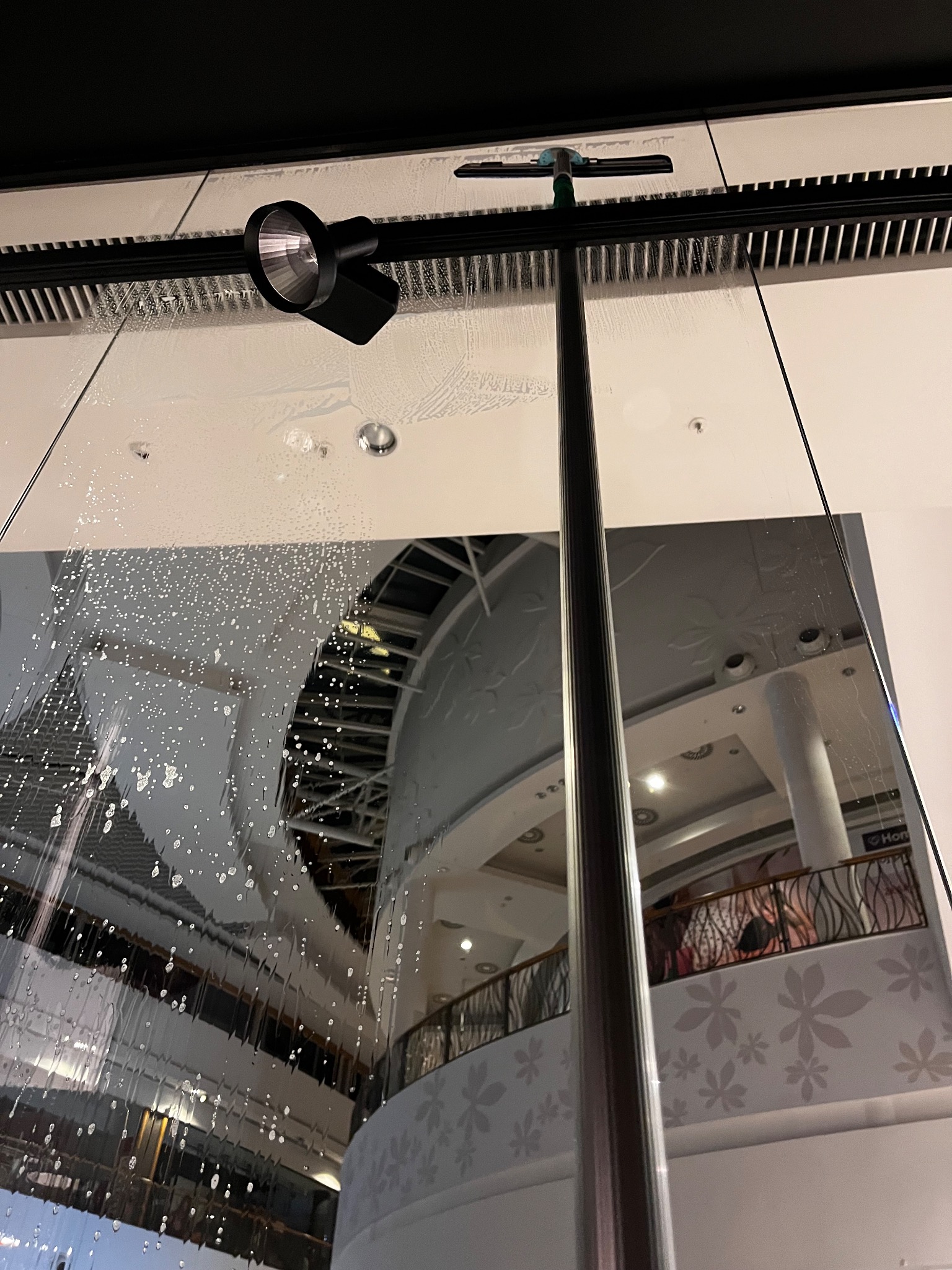 Mycie dużej, przeszklonej powierzchni wewnątrz centrum handlowego z użyciem myjki na długim kiju. Widoczne ślady wody i piany na szybie, odblask wnętrza centrum.