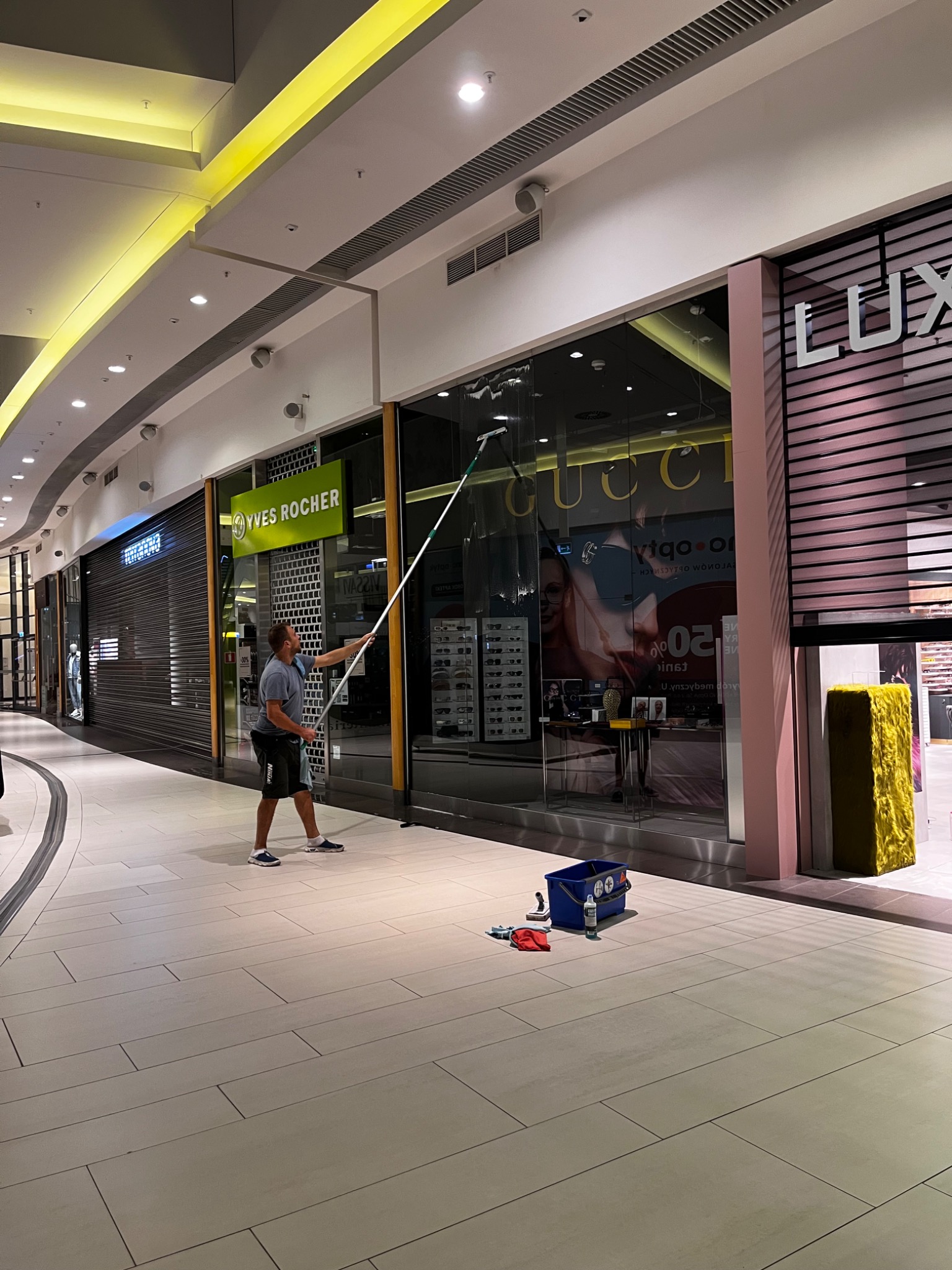 Pracownik w szarej koszulce czyści wysokie okno wystawowe sklepu Gucci w galerii handlowej za pomocą długiego mopa, obok stoi niebieskie wiadro z akcesoriami.