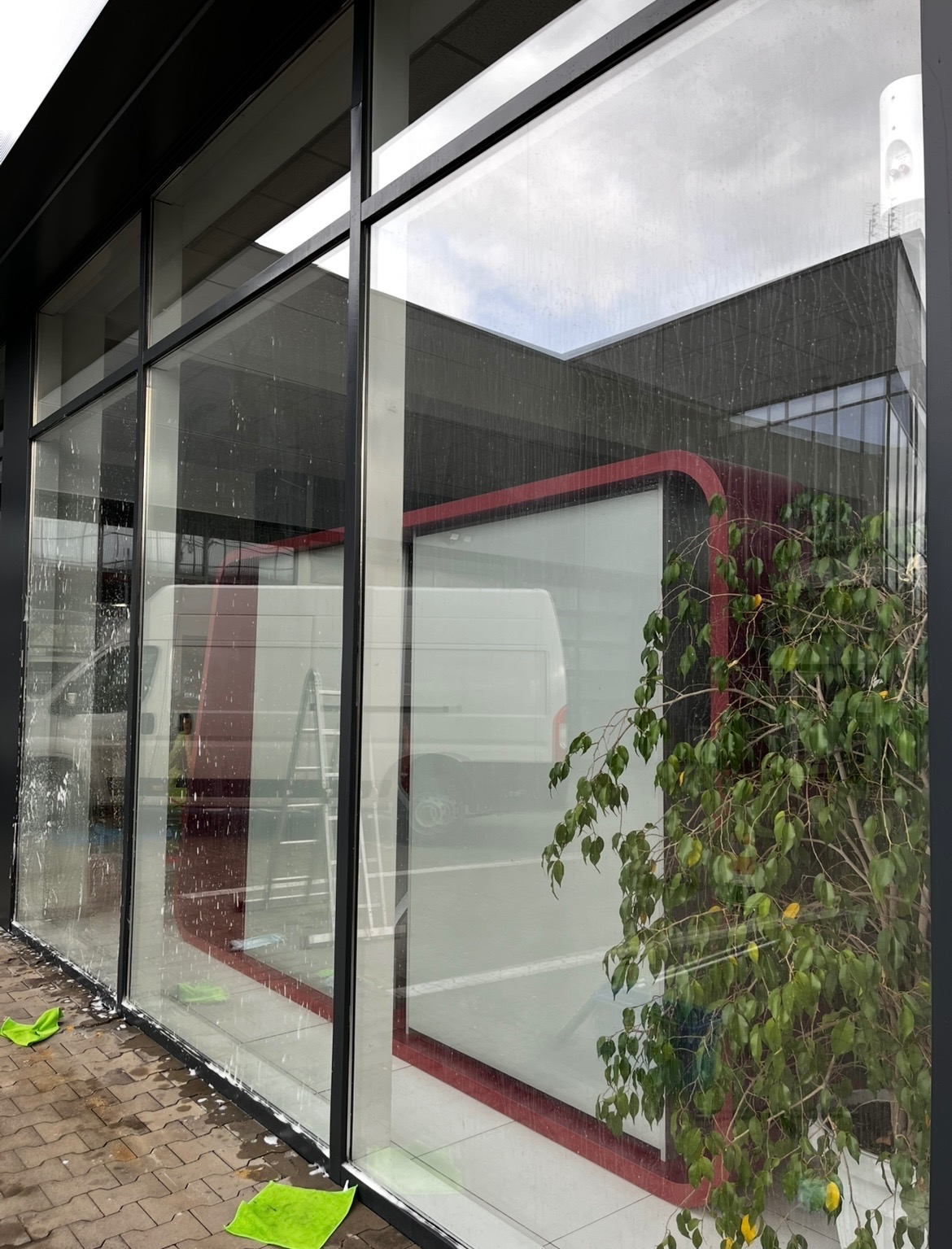 Mycie okien biurowych z widocznymi smugami i pianą, zielone ścierki na parapecie, drabina i samochód dostawczy w tle za szybą.