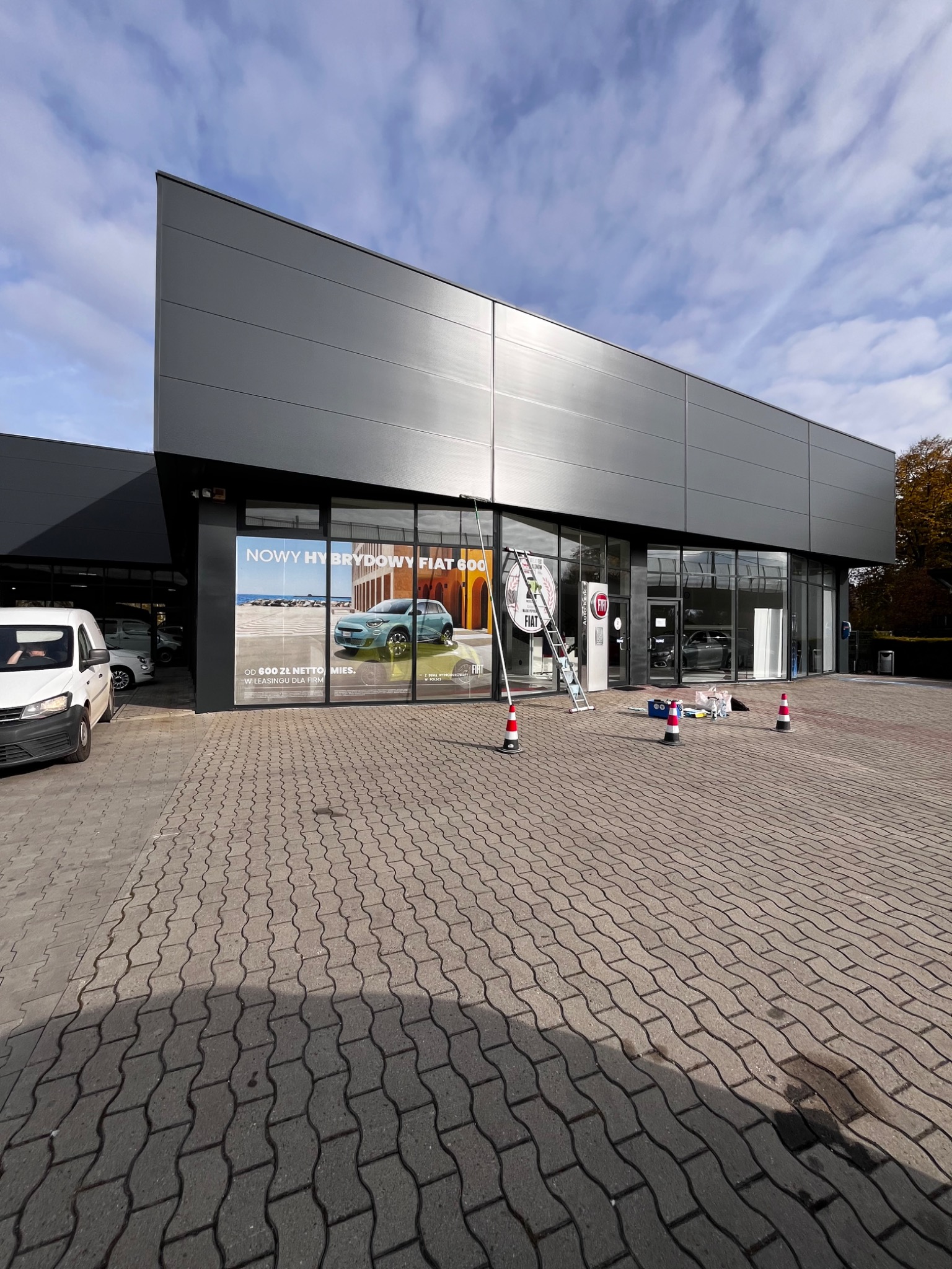 Mycie okien salonu samochodowego Fiat, widoczna drabina oparta o szybę, narzędzia i pachołki na chodniku.