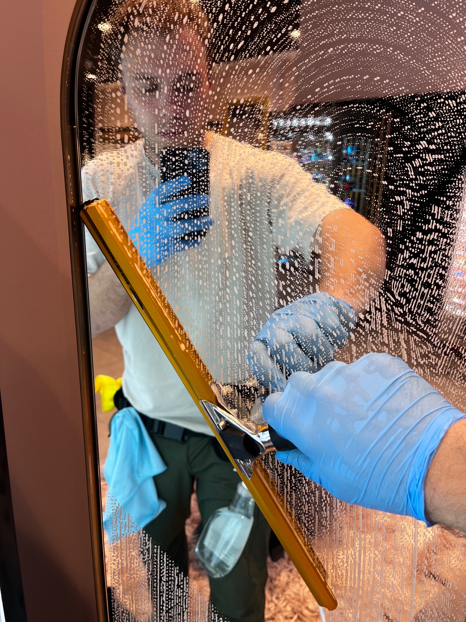 Osoba w niebieskich rękawicach myje lustro za pomocą ściągaczki z żółtą rączką, widoczne ślady piany i odbicie osoby w lustrze.