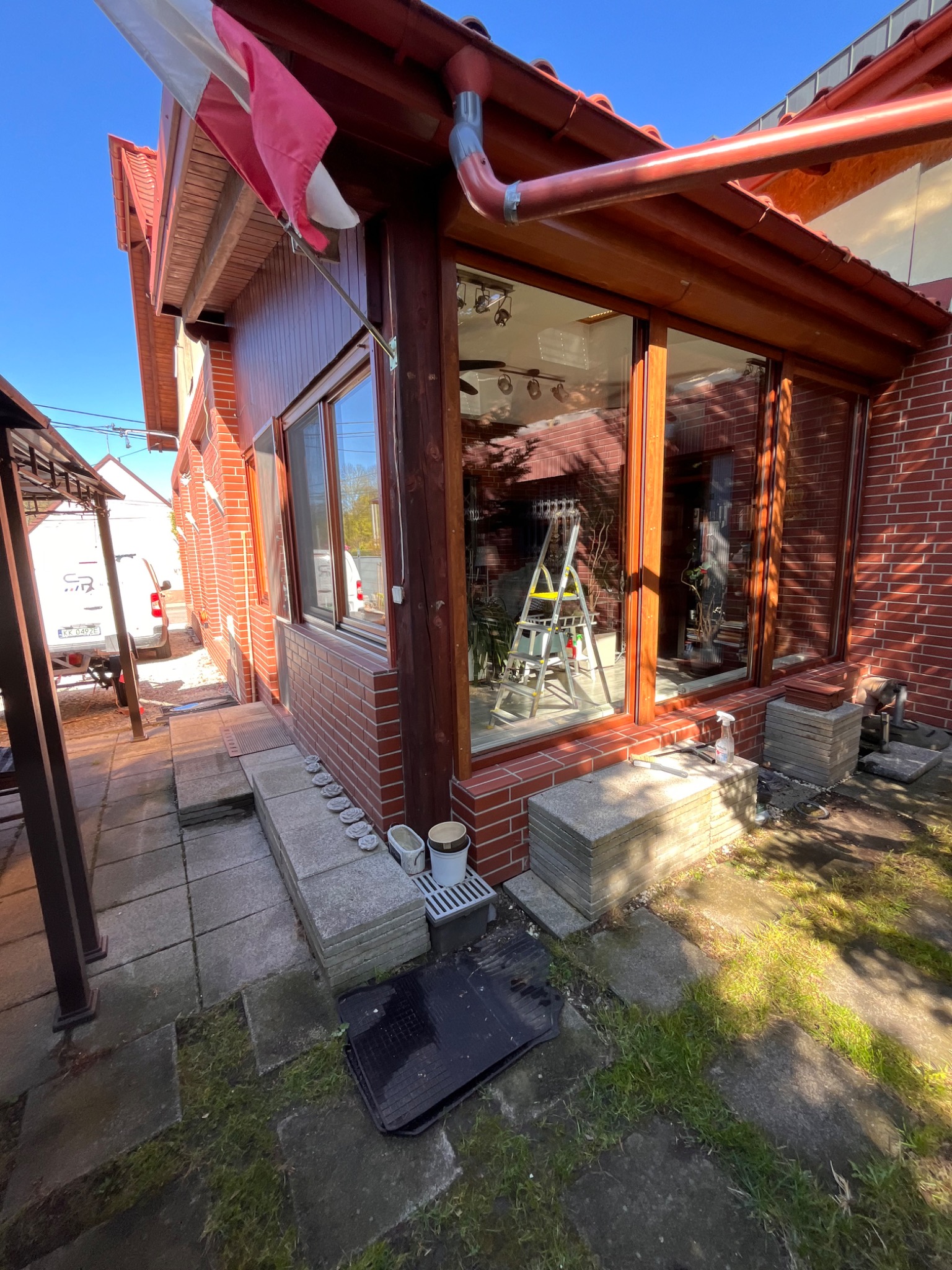 Zewnętrzny widok domu z czerwonej cegły z dużymi oknami, na których stoi aluminiowa drabina i butelka ze spryskiwaczem, sugerująca przygotowania do czyszczenia.