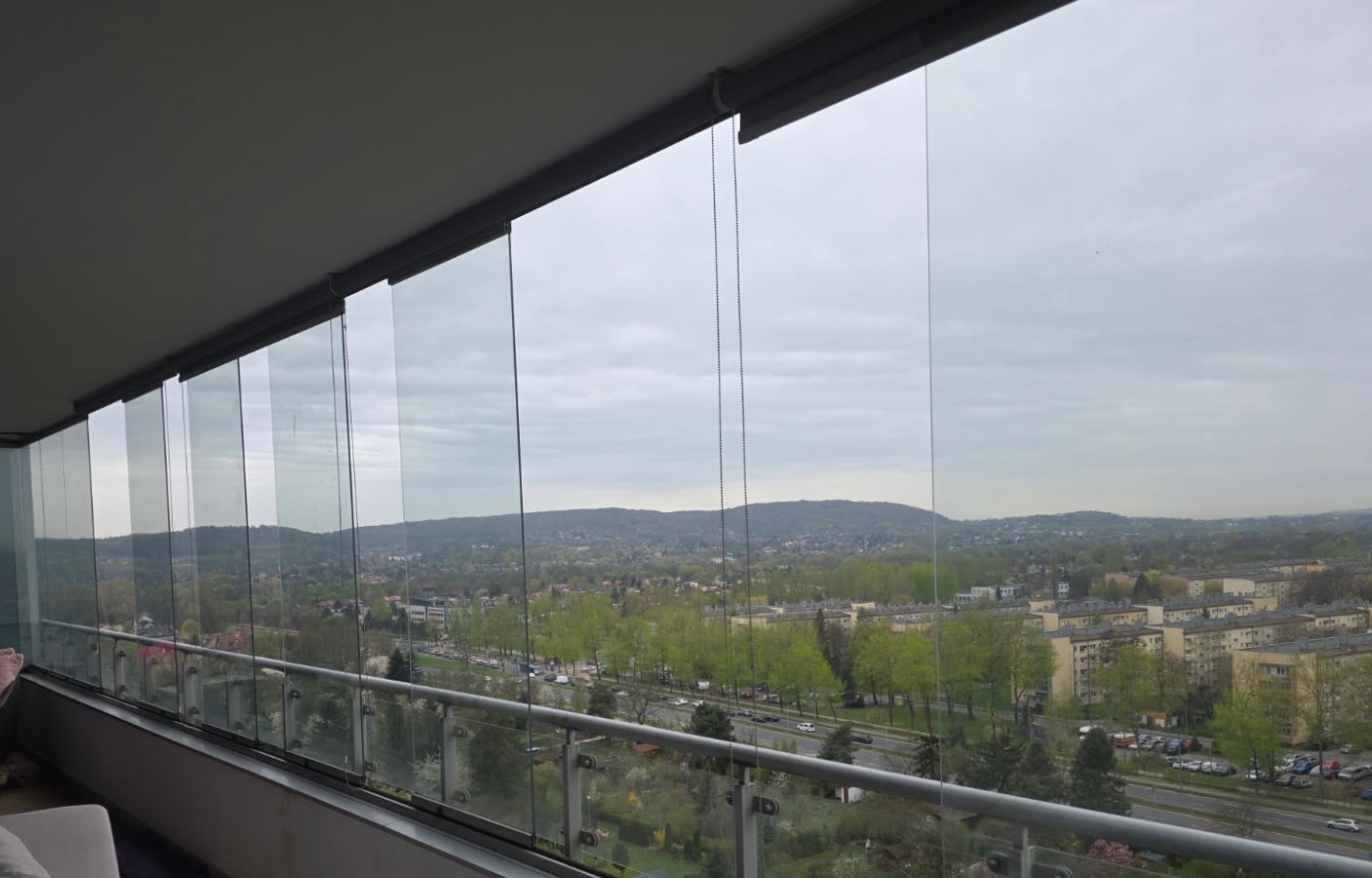 Zabudowany balkon z przesuwnymi szklanymi panelami, widok na miasto z zielenią i wzgórzami w tle, pochmurne niebo.