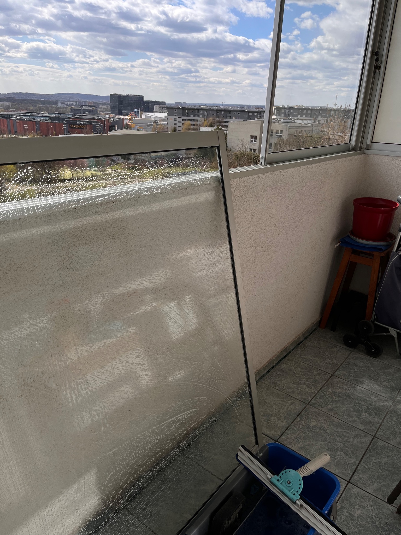 Okno balkonowe w trakcie czyszczenia, widoczne krople wody z detergentem, w tle panorama miasta, niebieskie wiadro i ściągaczka do szyb.