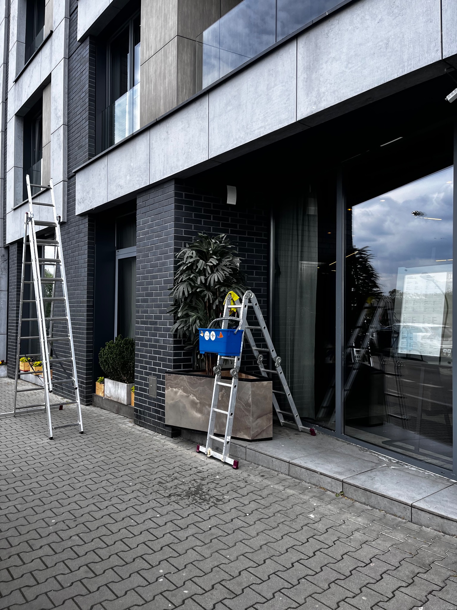 Dwie drabiny przy nowoczesnym budynku z przeszklonym wejściem. Jedna drabina z niebieskim pojemnikiem na środku, druga oparta o ścianę. Prawdopodobnie mycie okien.
