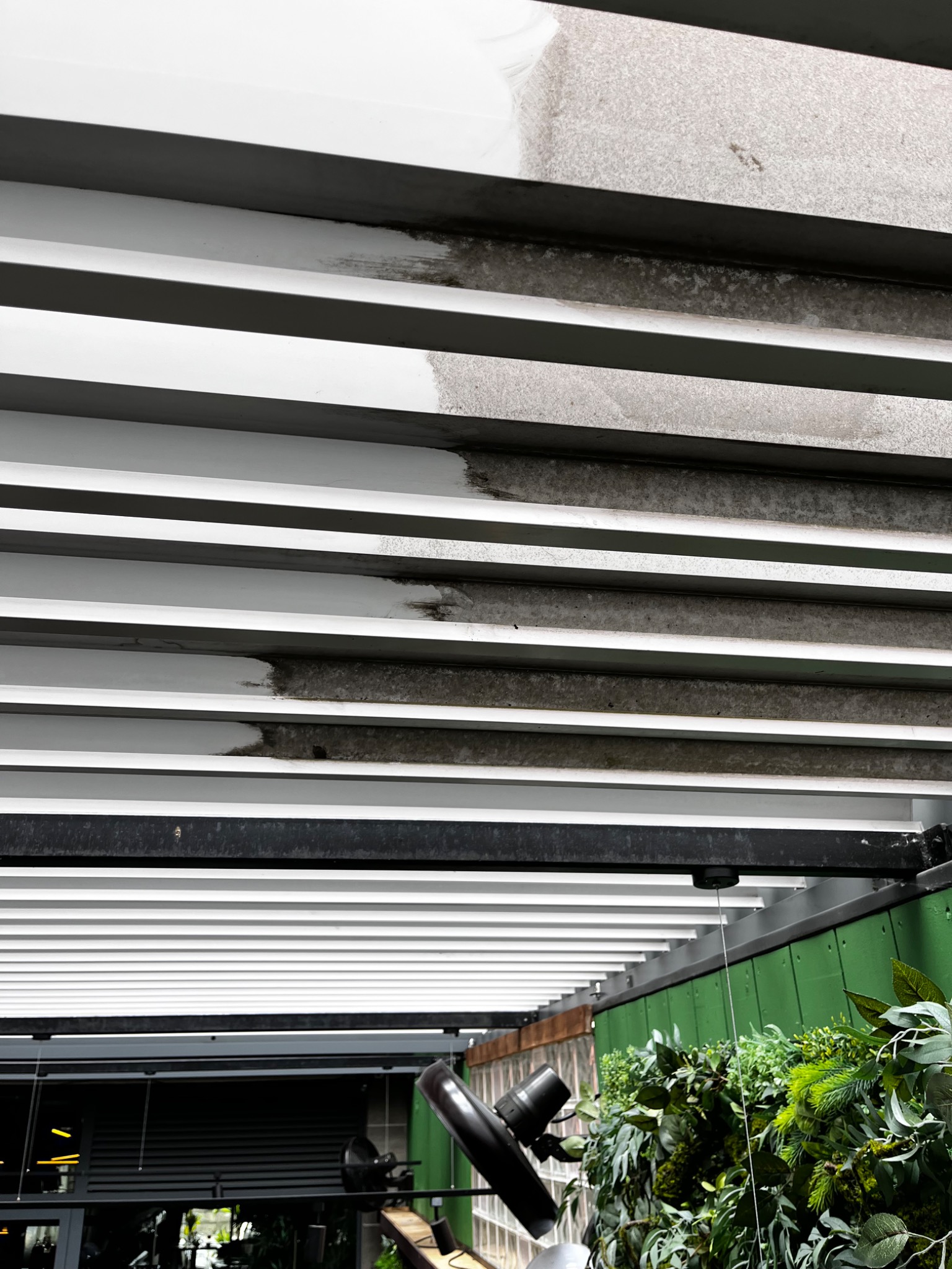 Szczegółowe ujęcie fragmentu czyszczonej, białej, poziomej żaluzji sufitowej, ukazujące różnicę między obszarem przed i po zabiegu. Widoczne zabrudzenia i świeżo umyta powierzchnia.