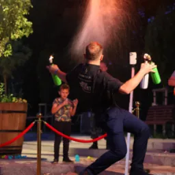 Barman w czarnej koszuli z logo firmy, stojący na obrotowej platformie, wykonuje pokaz z płomieniami i rozpryskującą się cieczą z butelek; w tle dziecko i dekoracje.
