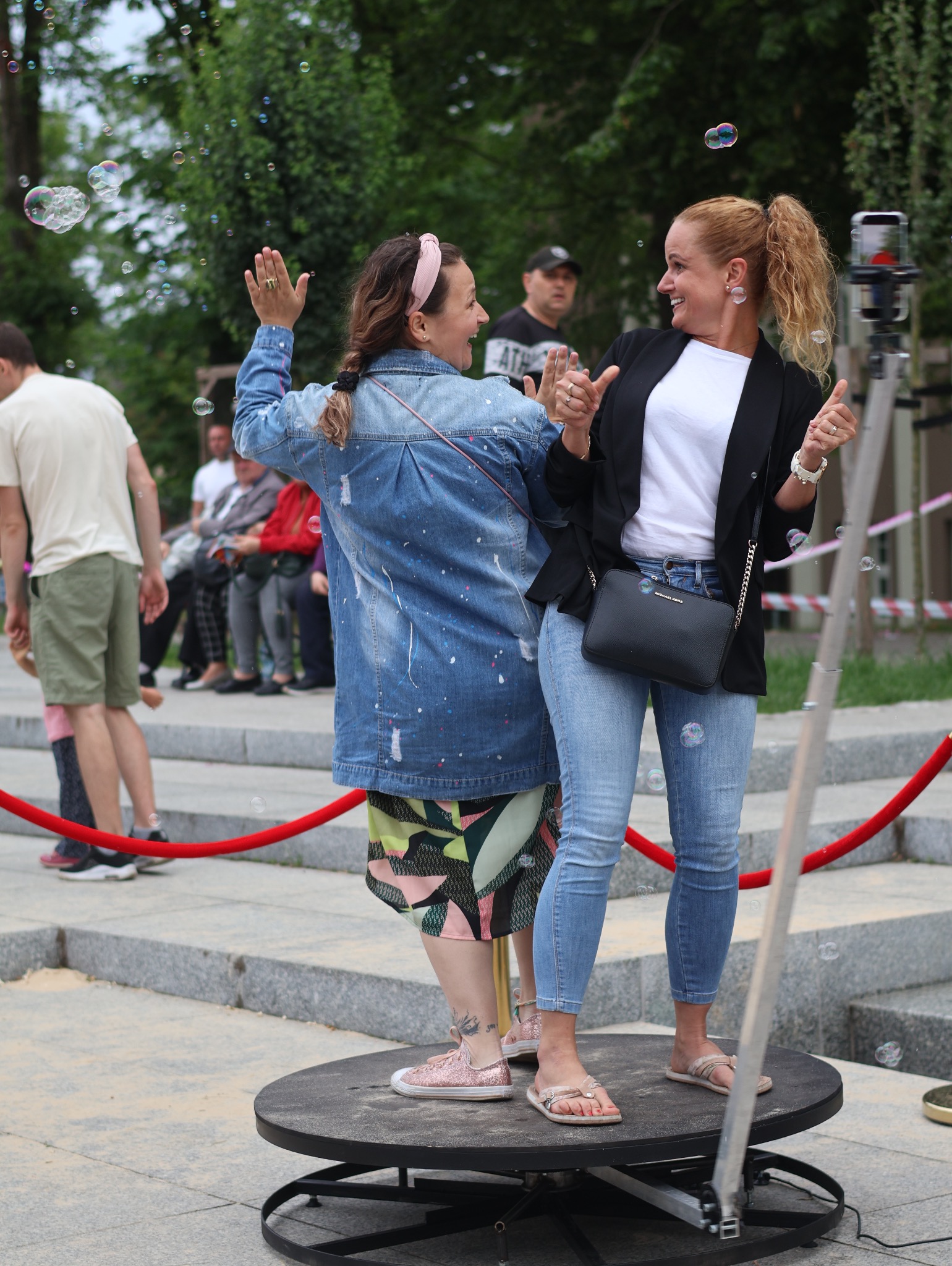 Dwie uśmiechnięte kobiety w otoczeniu baniek mydlanych, stojące na obrotowej platformie podczas sesji zdjęciowej.