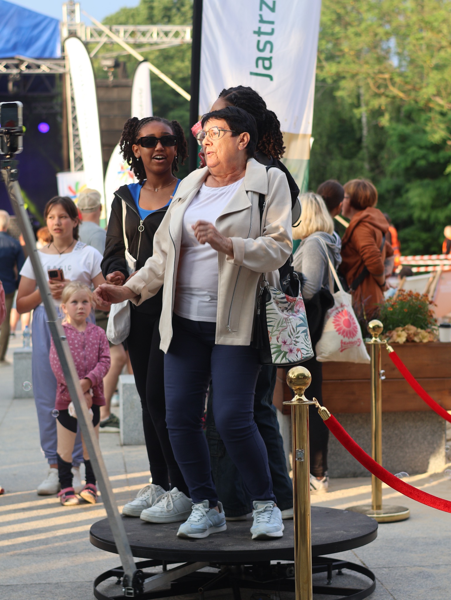 Trzy osoby pozują na obrotowej platformie fotobudki 360 stopni podczas imprezy plenerowej, widoczny smartfon na statywie, złote słupki z czerwonym sznurem i tłum ludzi w tle.