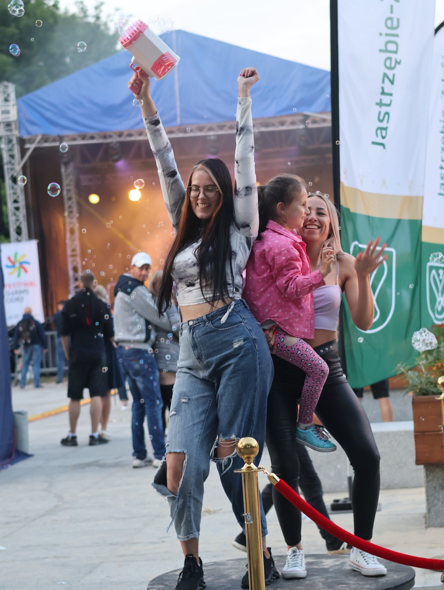 Uśmiechnięta kobieta w okularach trzyma w górze pistolet do baniek mydlanych na tle sceny z niebieskim zadaszeniem i bawiących się ludzi, w pobliżu stoją dwie inne kobiety i dziecko.