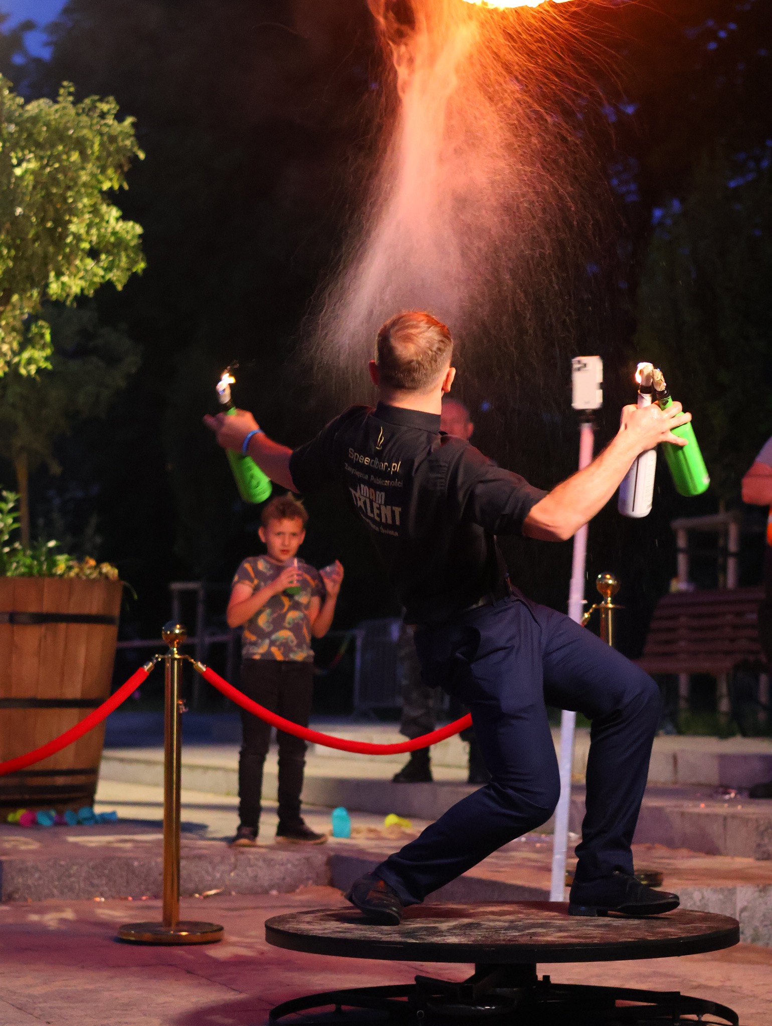 Barman w czarnej koszuli z logo firmy, stojący na obrotowej platformie, wykonuje pokaz z płomieniami i rozpryskującą się cieczą z butelek; w tle dziecko i dekoracje.