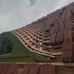Budowa dachu z nietypowym, łukowym oknem dachowym; przygotowane do położenia dachówki ceramicznej, widoczne drewniane łaty i kontrłaty oraz pakiety dachówek czekające na montaż.