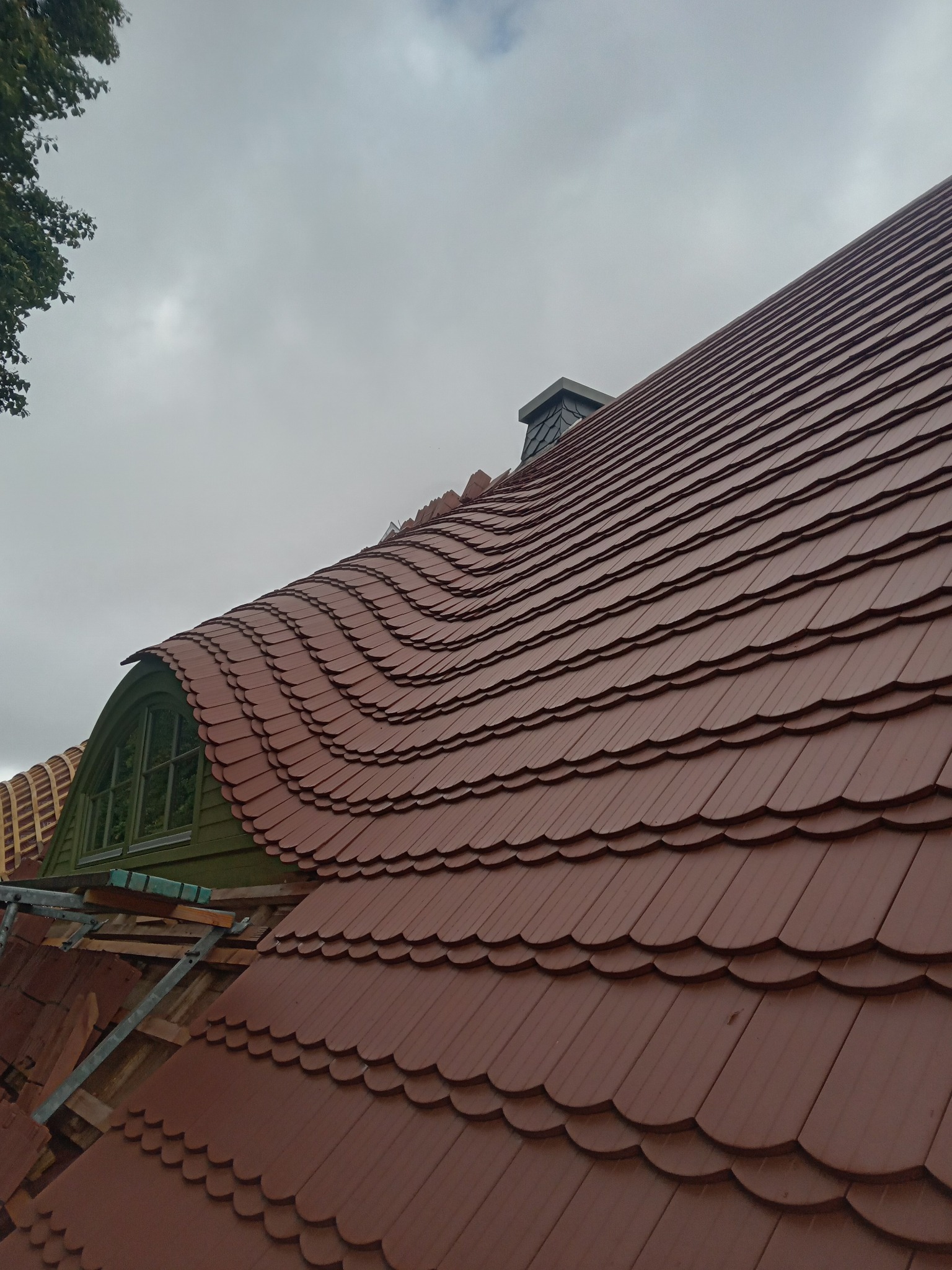 Fragment dachu z nową, brązową dachówką karpiówką w trakcie układania, widoczne rusztowanie i okno dachowe w kształcie łuku.