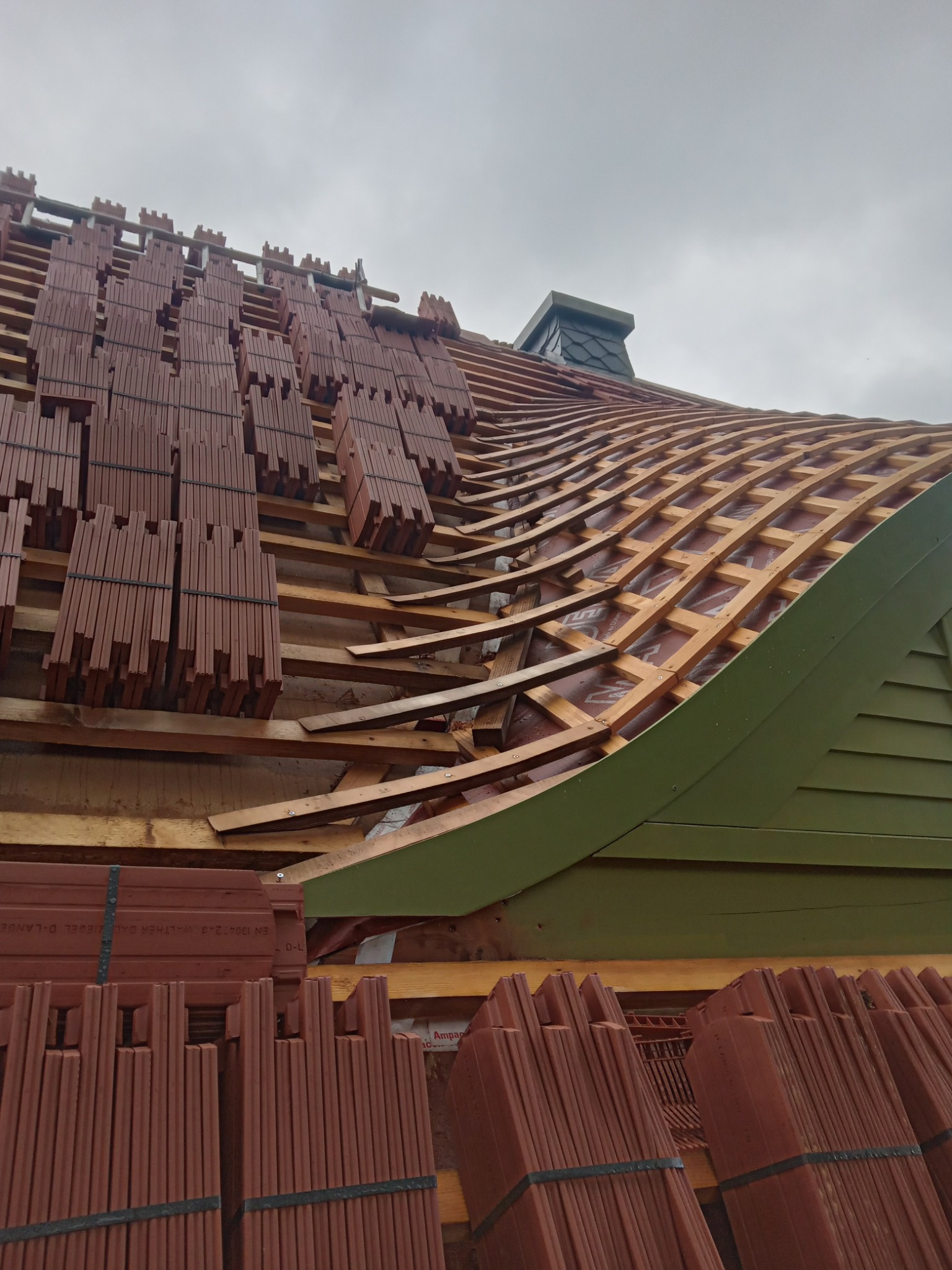 Układanie dachówki ceramicznej na łukowatym dachu z widoczną konstrukcją drewnianą i kominem krytym łupkiem, z paletami dachówek gotowych do montażu.