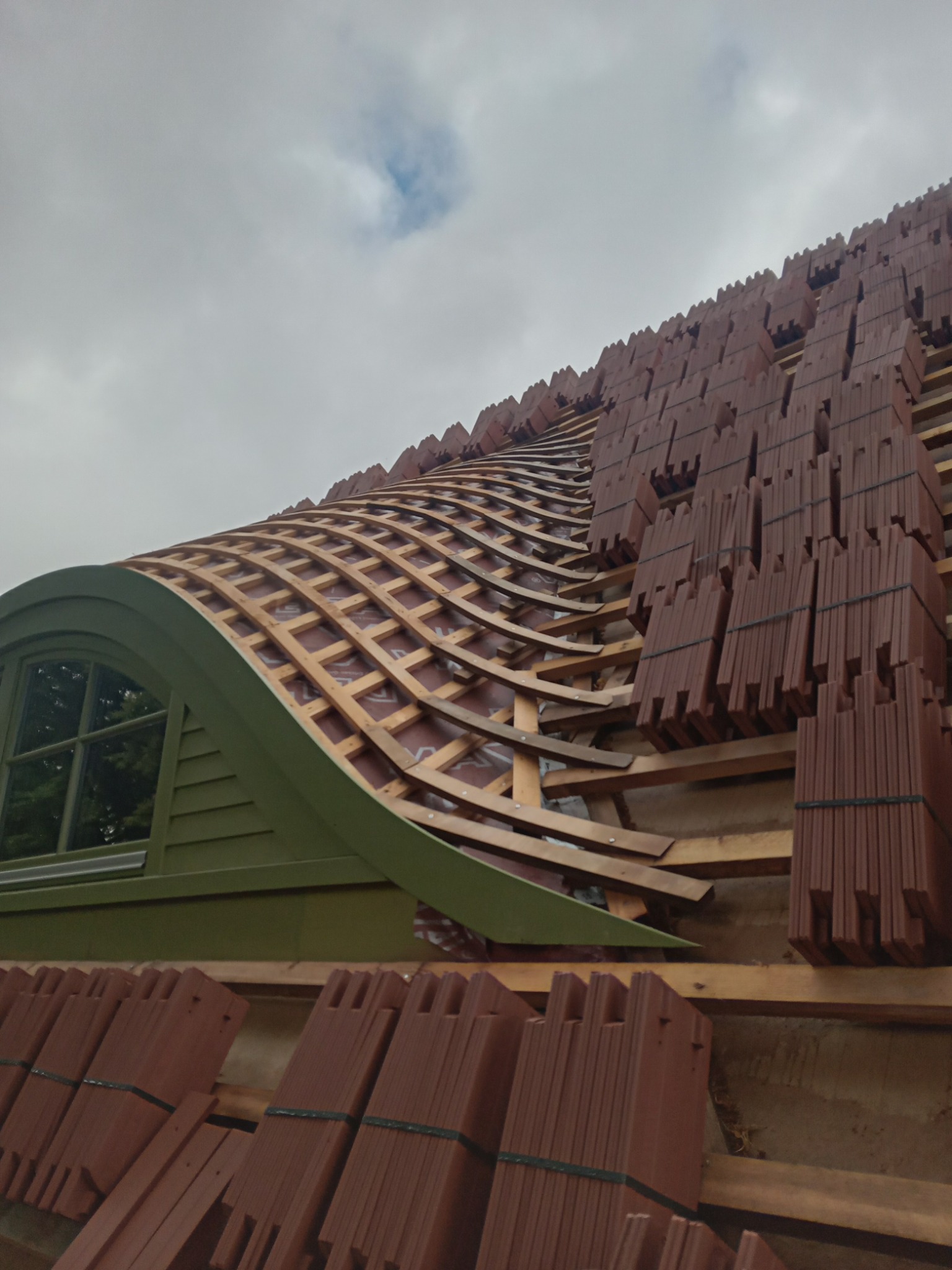 Budowa dachu z nietypowym, łukowym oknem dachowym; przygotowane do położenia dachówki ceramicznej, widoczne drewniane łaty i kontrłaty oraz pakiety dachówek czekające na montaż.