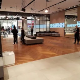 Wnętrze nowoczesnego sklepu z drewnianą podłogą ułożoną w jodełkę, szarymi sofami i ścianami z wyświetlaczami prezentującymi produkty. Widoczni klienci i pracownicy.