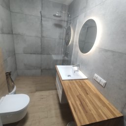 LEMARTIN - Nowoczesna łazienka z szarymi płytkami imitującymi beton, drewnianym blatem pod umywalką, okrągłym lustrem z podświetleniem LED i kabiną prysznicową typu walk-in z widoczną deszczownicą.