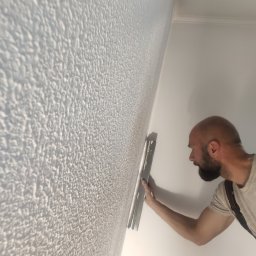 LEMARTIN - Aplikacja dekoracyjnej struktury na ścianie przez wykonawcę z użyciem pacy. Widoczna faktura ściany i narzędzie pracy.