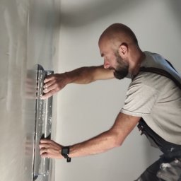 LEMARTIN - Mężczyzna w pracy, gładzi ścianę szpachelką. Widoczny fragment wygładzonej powierzchni, narzędzia przy pasie i profil robotnika.