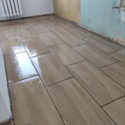 Ułożone, prostokątne płytki podłogowe imitujące drewno, z ciemną fugą. W tle fragment ściany w kolorze beżowym i błękitnym, oraz biały grzejnik.