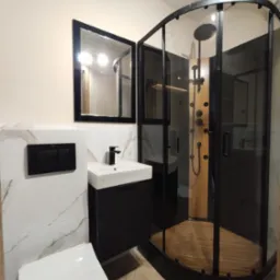 Nowoczesna łazienka po remoncie: kabina prysznicowa z drewnianym panelem, umywalka z czarną szafką i lustro w czarnej ramie. Białe płytki imitujące marmur.