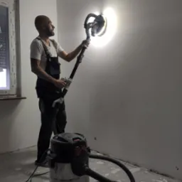 Szpachlowanie ściany szlifierką z odkurzaczem przez mężczyznę w kombinezonie roboczym. Widoczne okno z roletami i fragment podłogi w trakcie remontu.