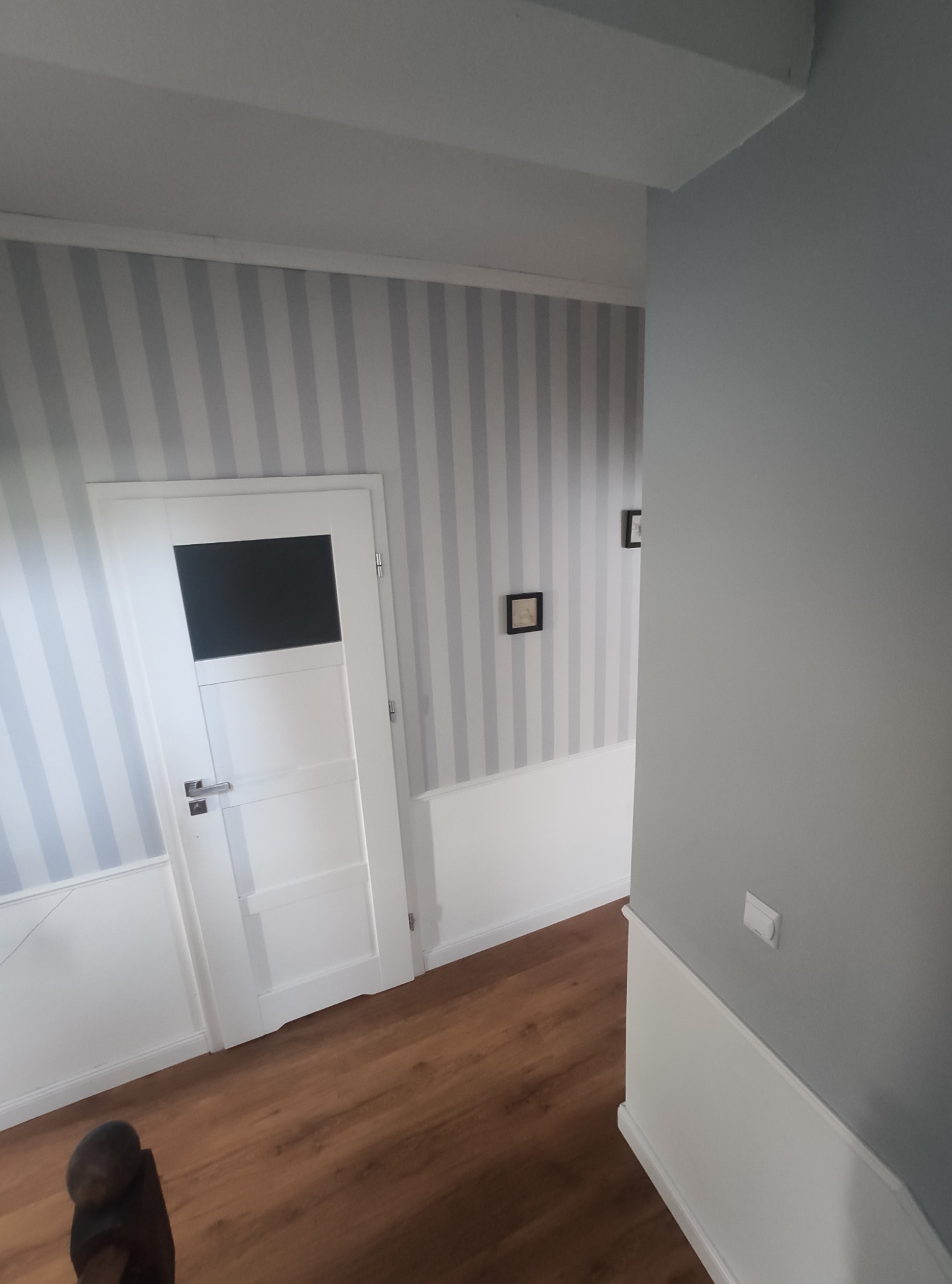 Korytarz po remoncie: ściany w szaro-białe pasy, biała stolarka drzwiowa z przeszkleniem, drewnopodobna podłoga i szara ściana boczna.