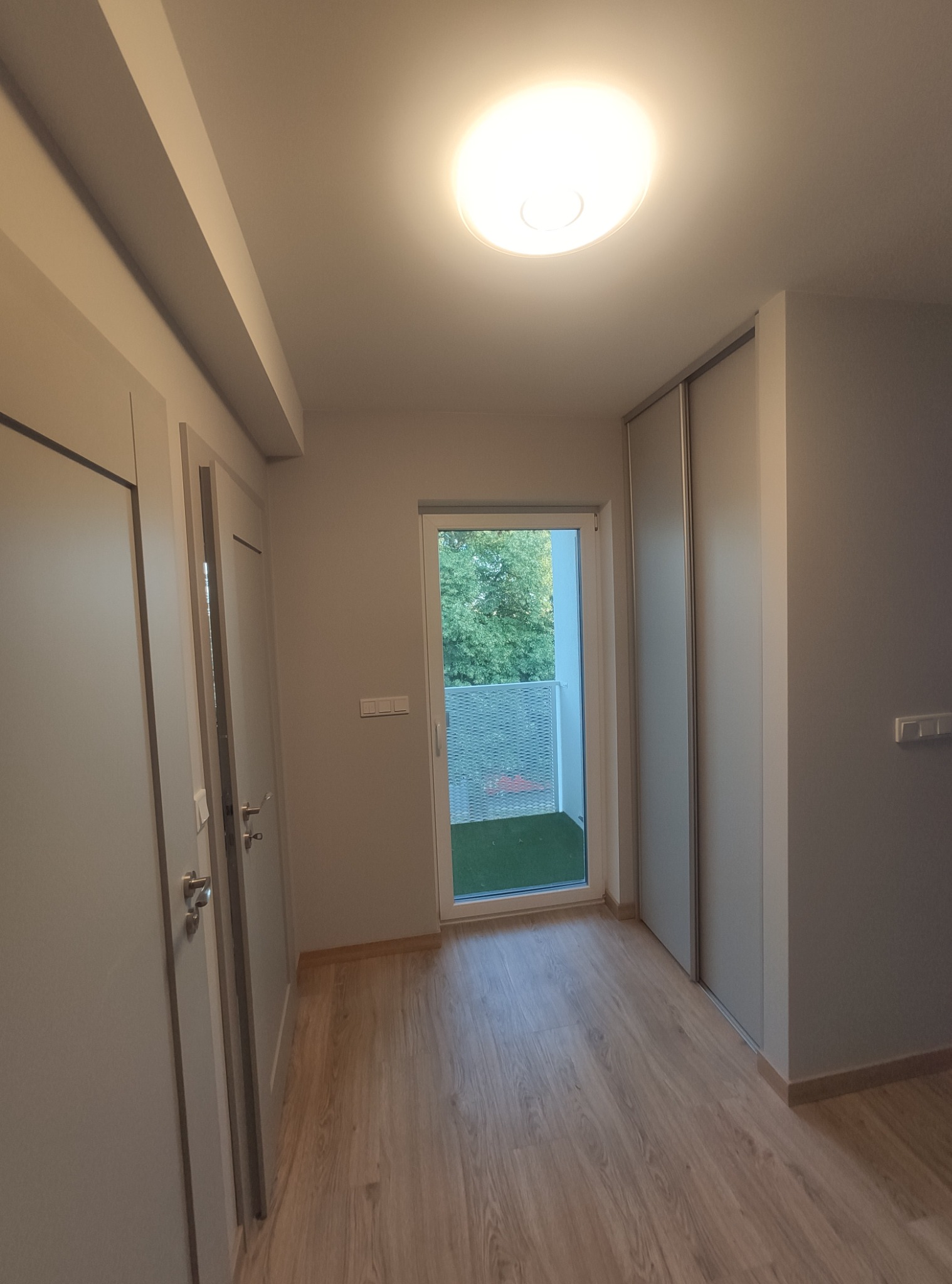 Korytarz po remoncie z drewnopodobną podłogą, szarą zabudową szafy przesuwnej i wyjściem na balkon.