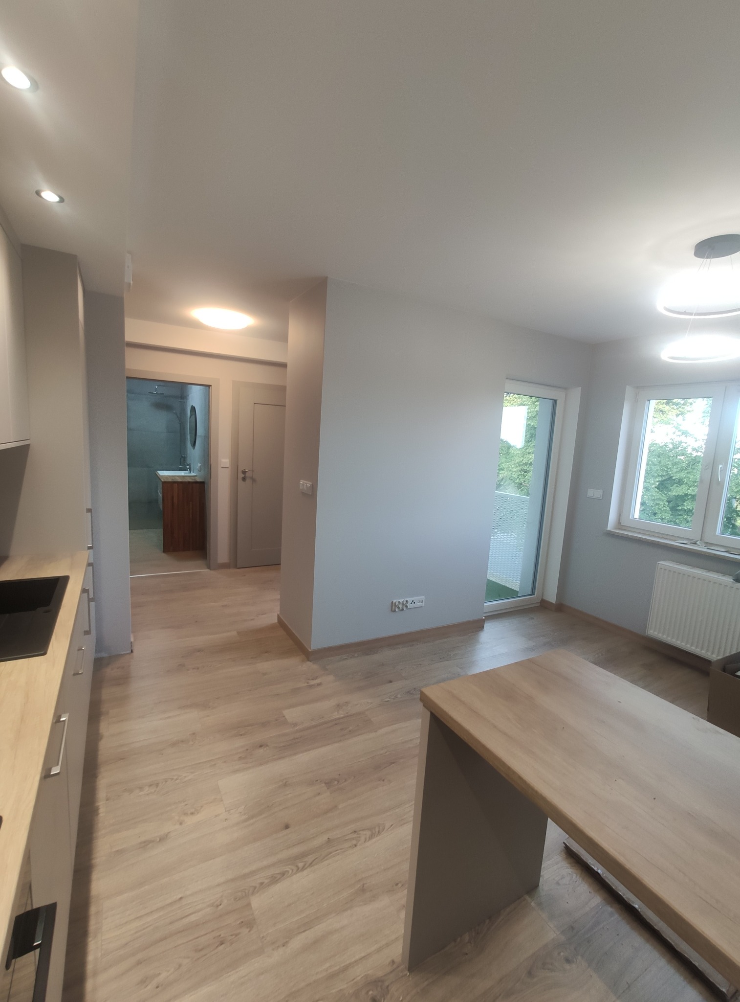 Jasne, przestronne wnętrze mieszkania po remoncie, z widoczną kuchnią, salonem z balkonem i łazienką w tle, wykończone w odcieniach szarości i drewna.