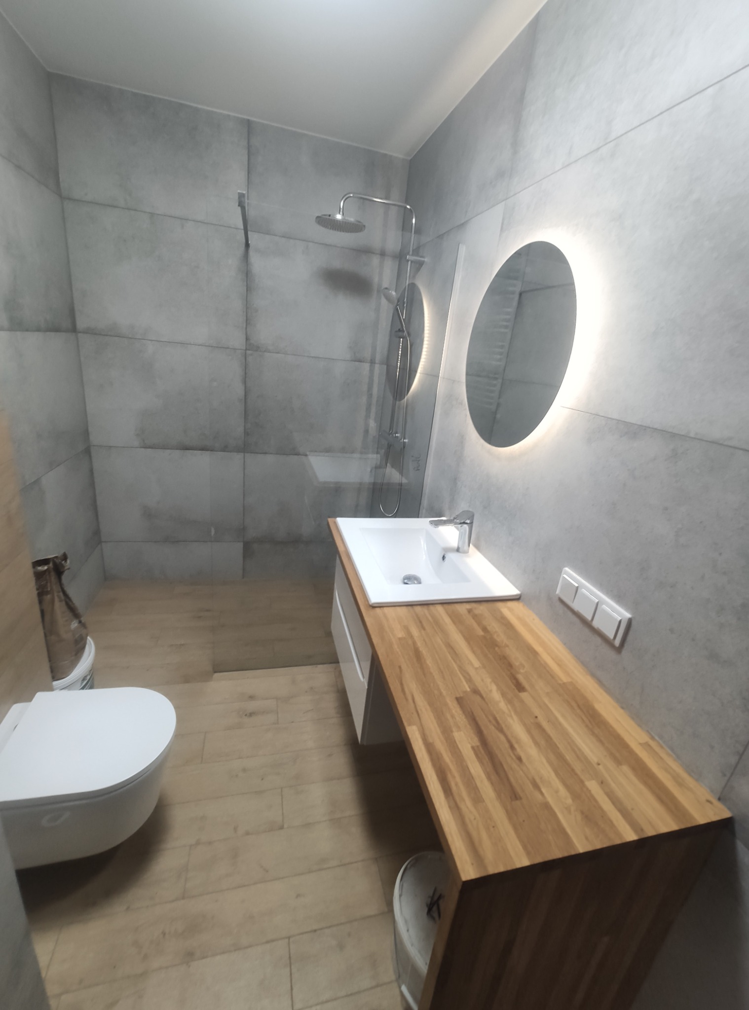 Nowoczesna łazienka z szarymi płytkami imitującymi beton, drewnianym blatem pod umywalką, okrągłym lustrem z podświetleniem LED i kabiną prysznicową typu walk-in z widoczną deszczownicą.