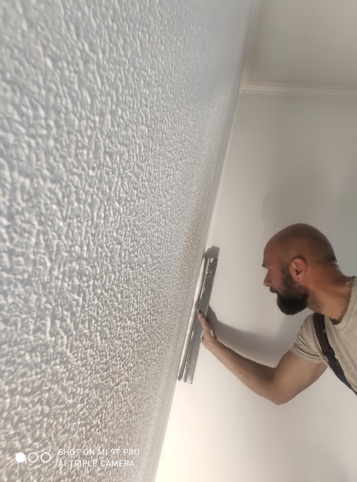 Aplikacja dekoracyjnej struktury na ścianie przez wykonawcę z użyciem pacy. Widoczna faktura ściany i narzędzie pracy.