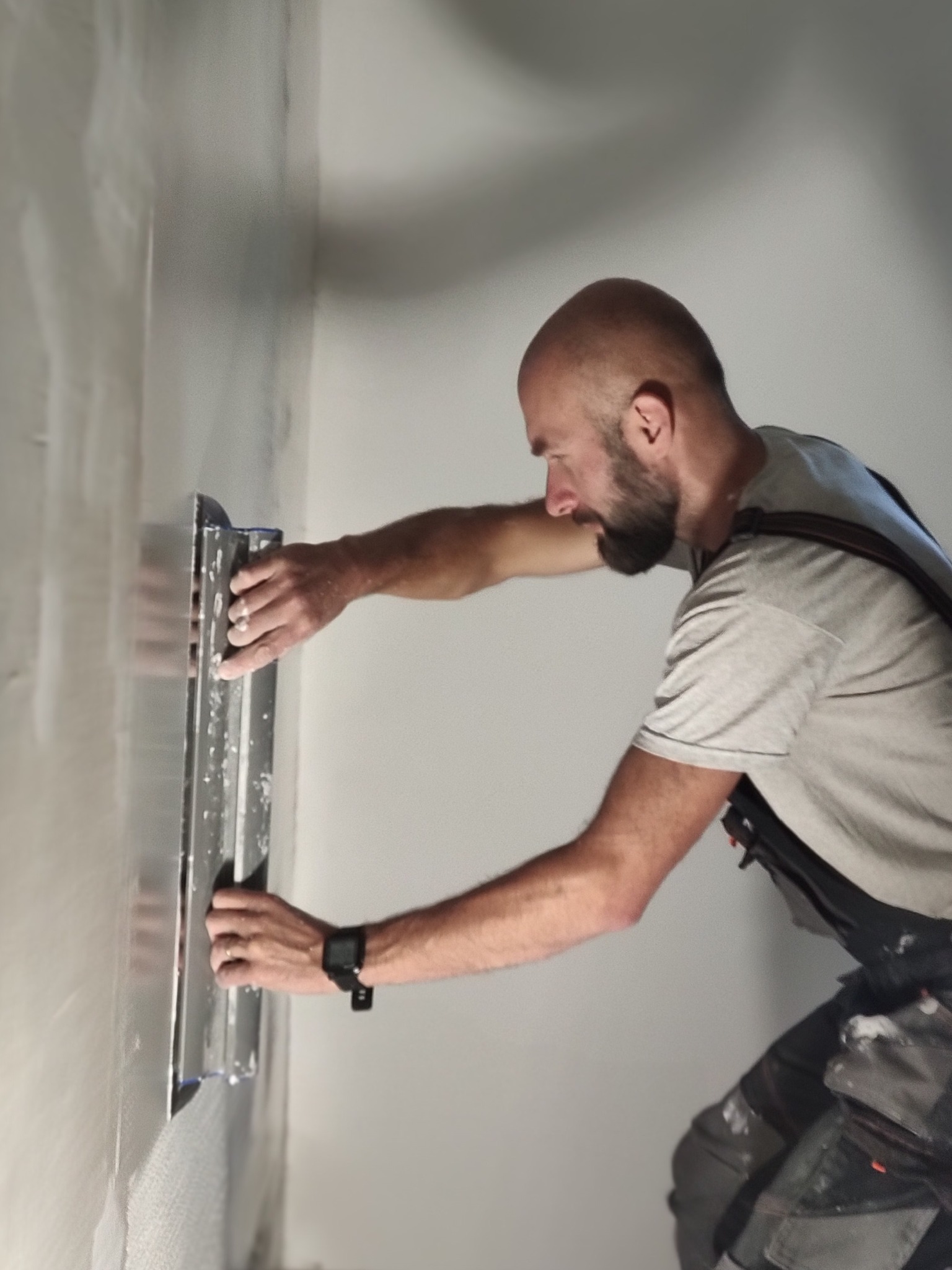 Mężczyzna w pracy, gładzi ścianę szpachelką. Widoczny fragment wygładzonej powierzchni, narzędzia przy pasie i profil robotnika.