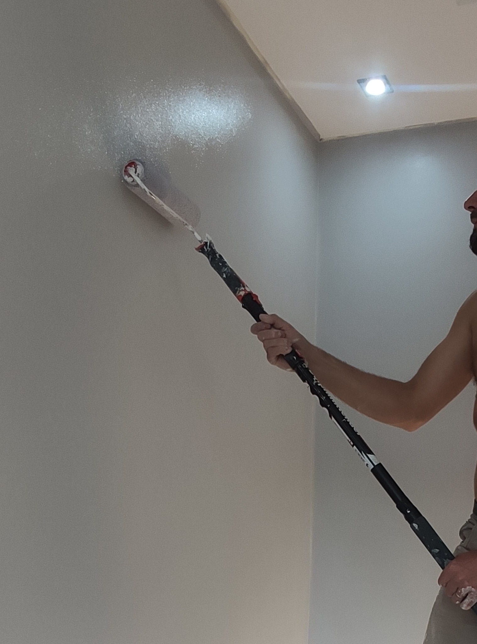 Mężczyzna maluje ścianę wałkiem na długim kiju, widoczny fragment twarzy i tors, oświetlenie sufitowe, szara ściana z efektem świeżego malowania.